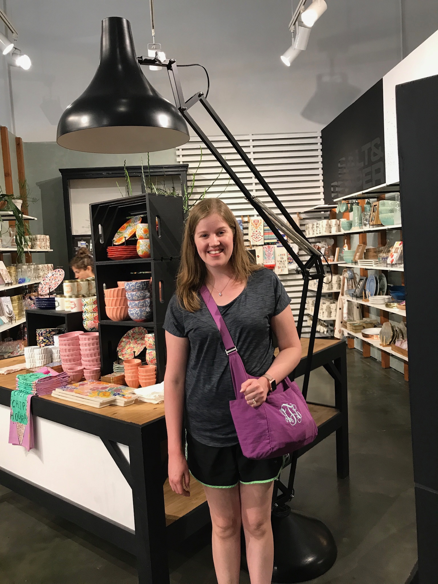 A young woman stands and smiles in a store with shelves of colorful dishes and ceramics. She is wearing a gray shirt, black shorts, and a purple shoulder bag.