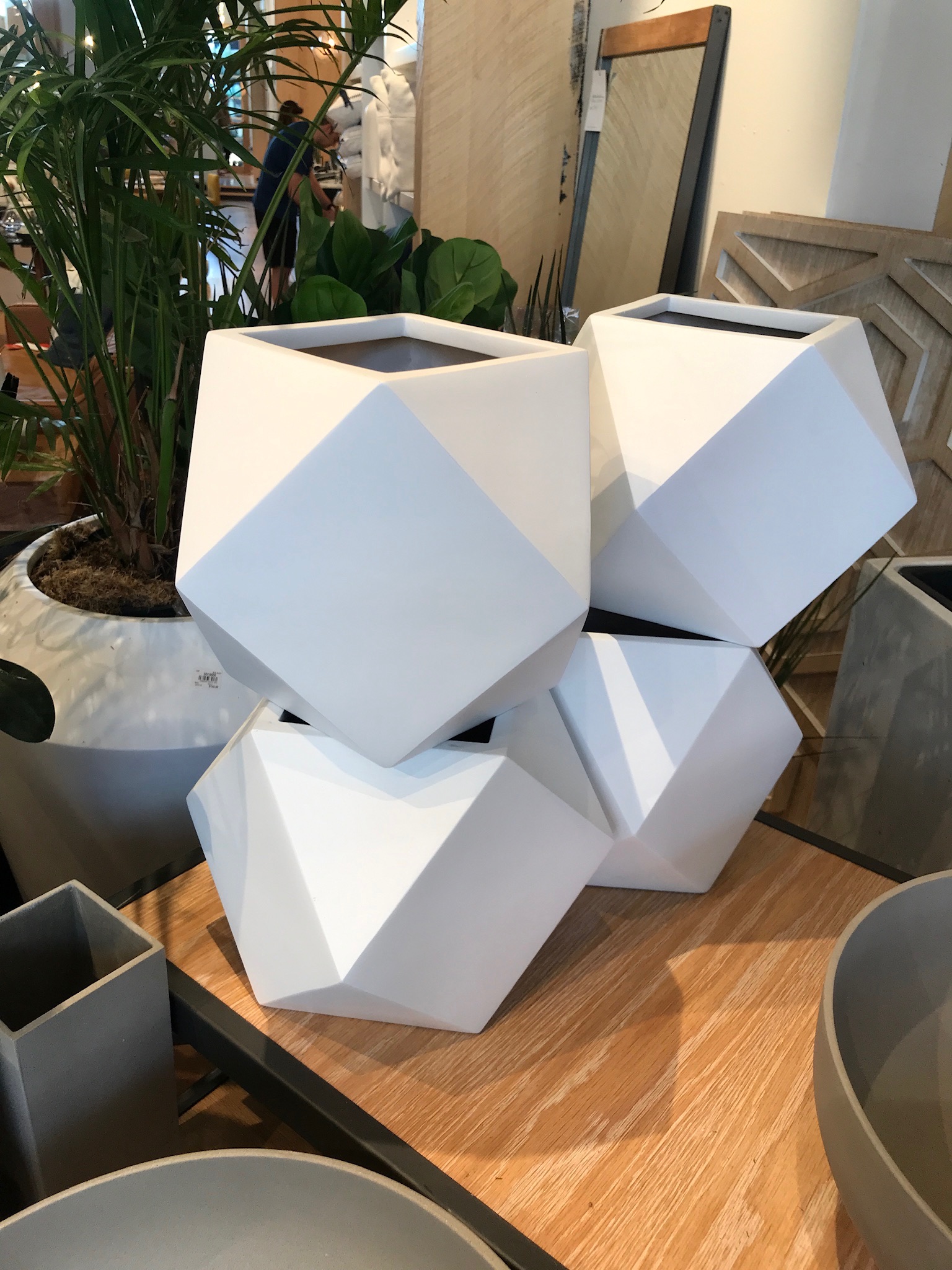 Four white geometric planters with faceted surfaces are stacked on a wooden table in a store, surrounded by other planters and decor items.