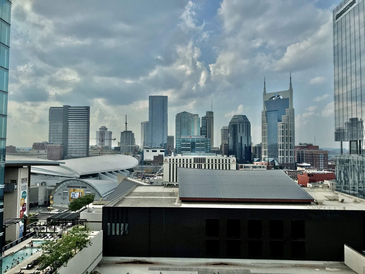 Nashville's city skyline showcases modern skyscrapers, highlighted by a prominent building with twin spires under a cloudy sky. In the foreground, rooftops and a partially visible pool buzzing with people add to the vibrant urban scene.
