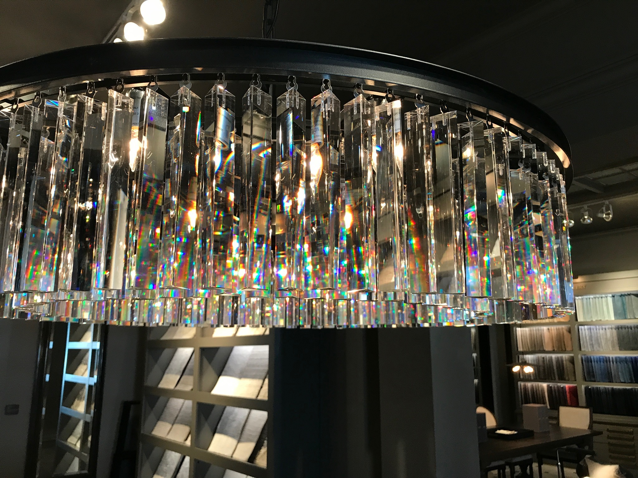 A close-up of a circular chandelier with hanging crystal prisms, lit bulbs, and rainbow light reflections, in a modern showroom with fabric samples in the background.