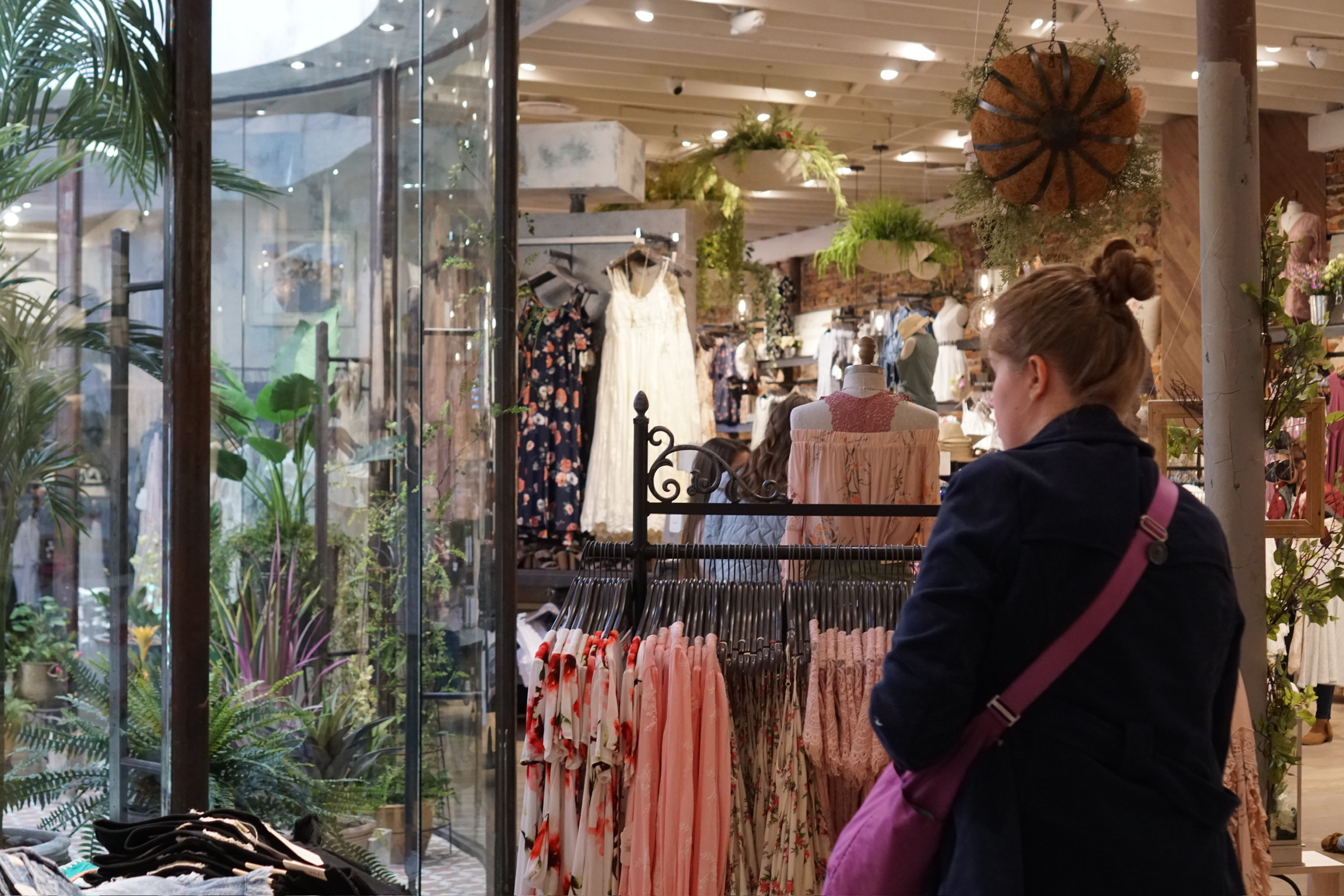 A woman wearing a navy coat and pink shoulder bag browses clothing racks in a boutique with hanging plants and dresses on display.