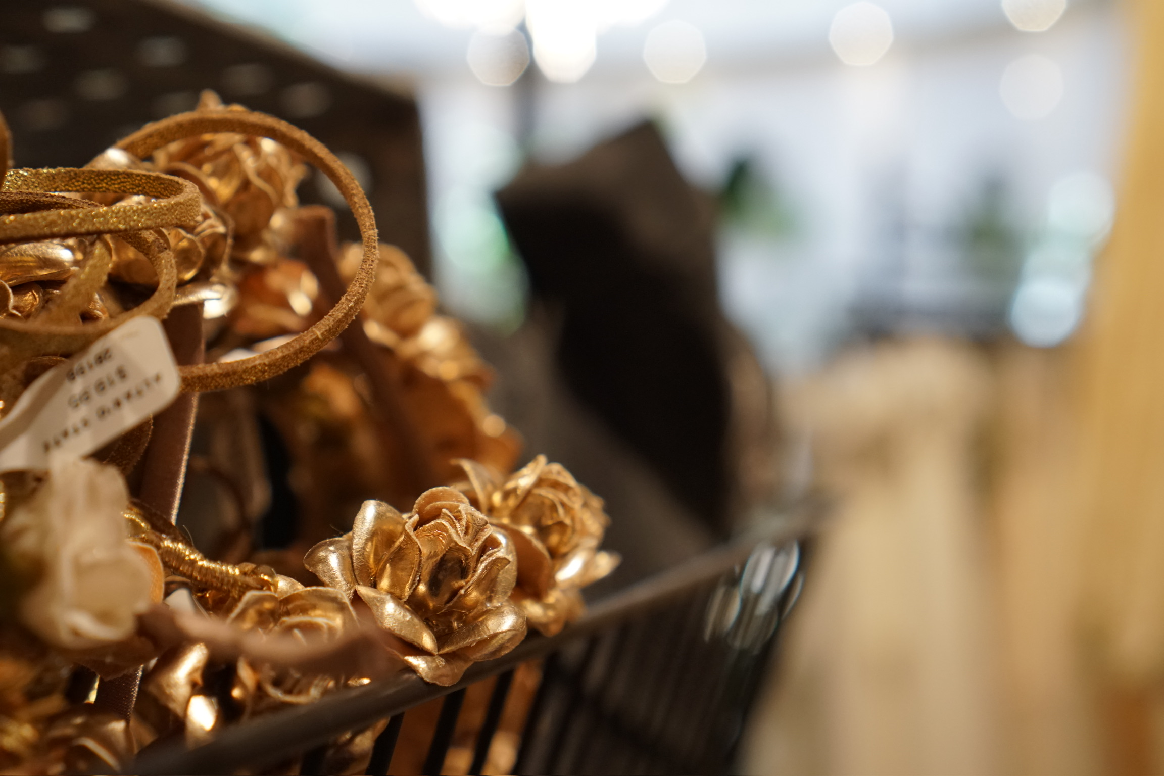 A close-up of a wire basket containing metallic gold floral headbands, with a blurred background of a shop interior.