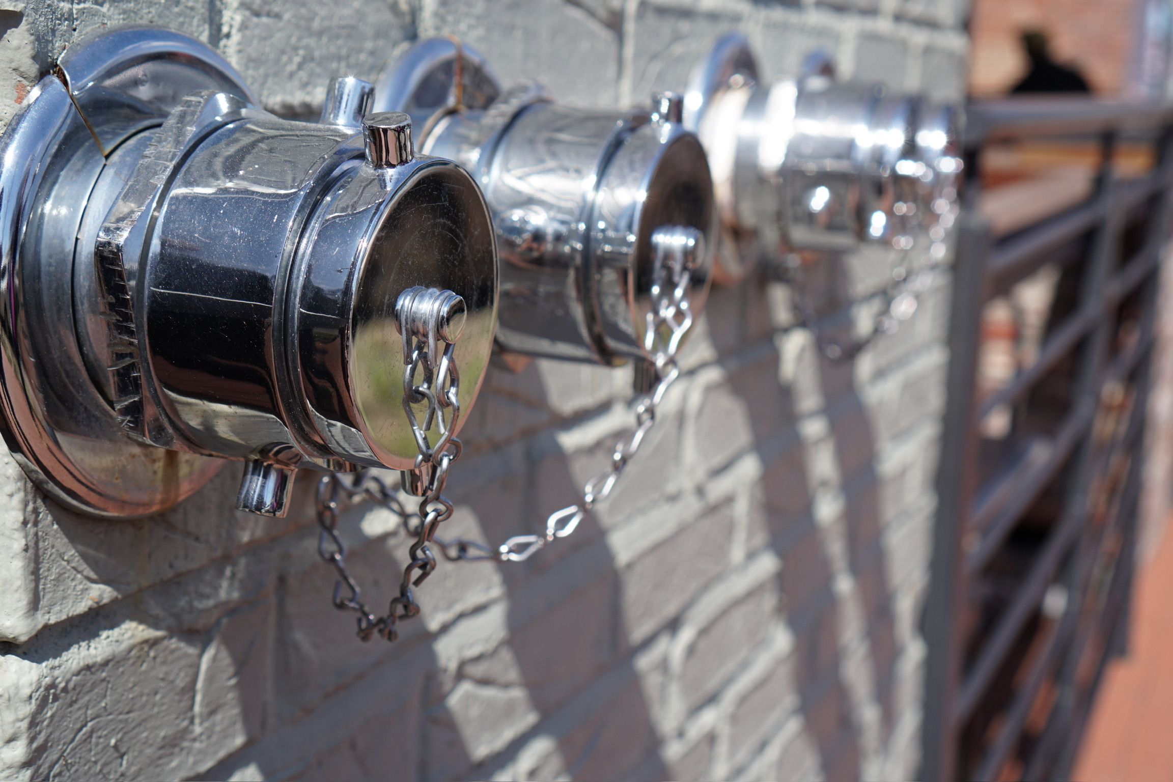 Close-up of three metal fire department connections with chains attached, mounted on a brick wall in an outdoor setting.