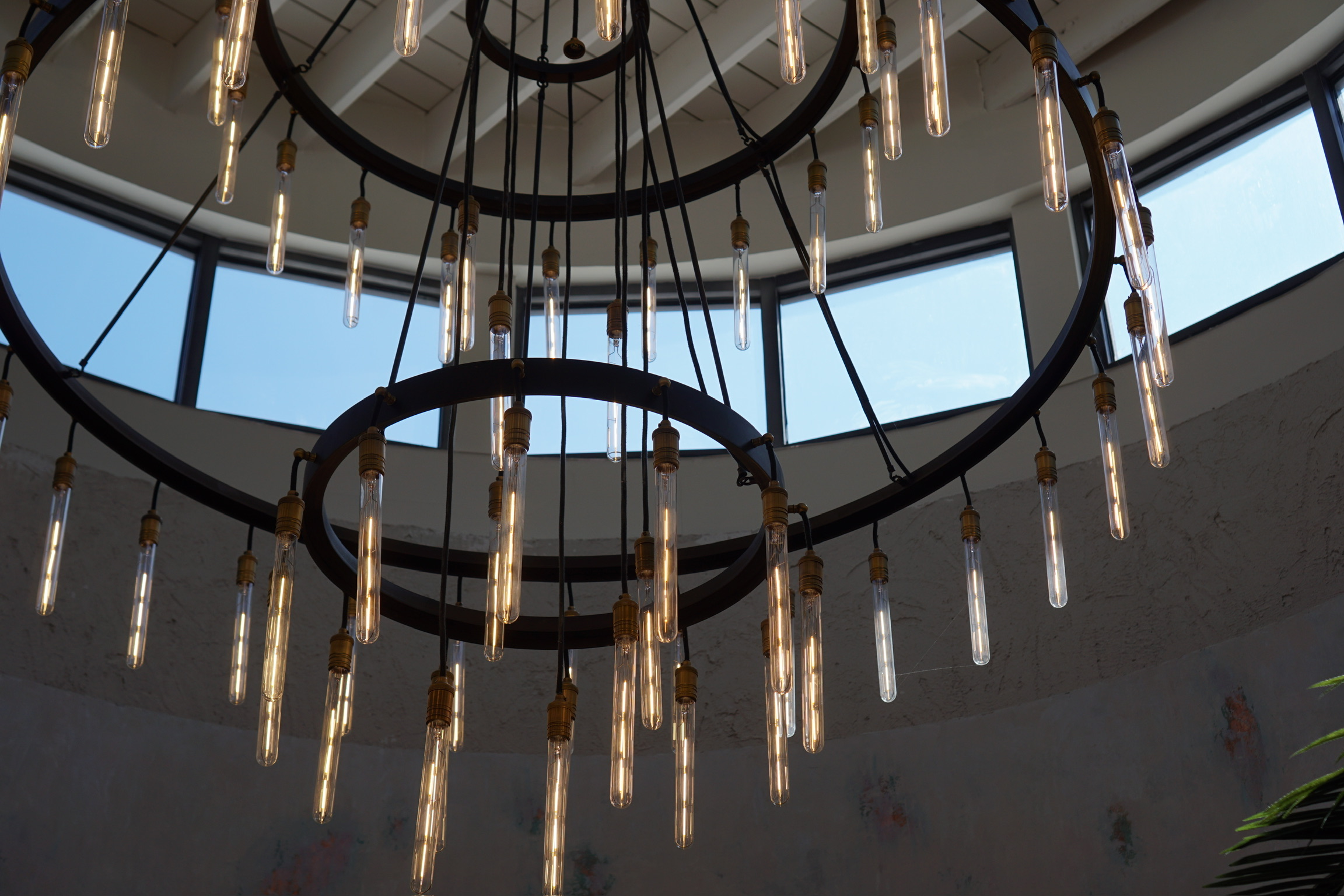Large circular chandelier with multiple exposed light bulbs hangs from a high ceiling beneath a round skylight with blue sky visible.