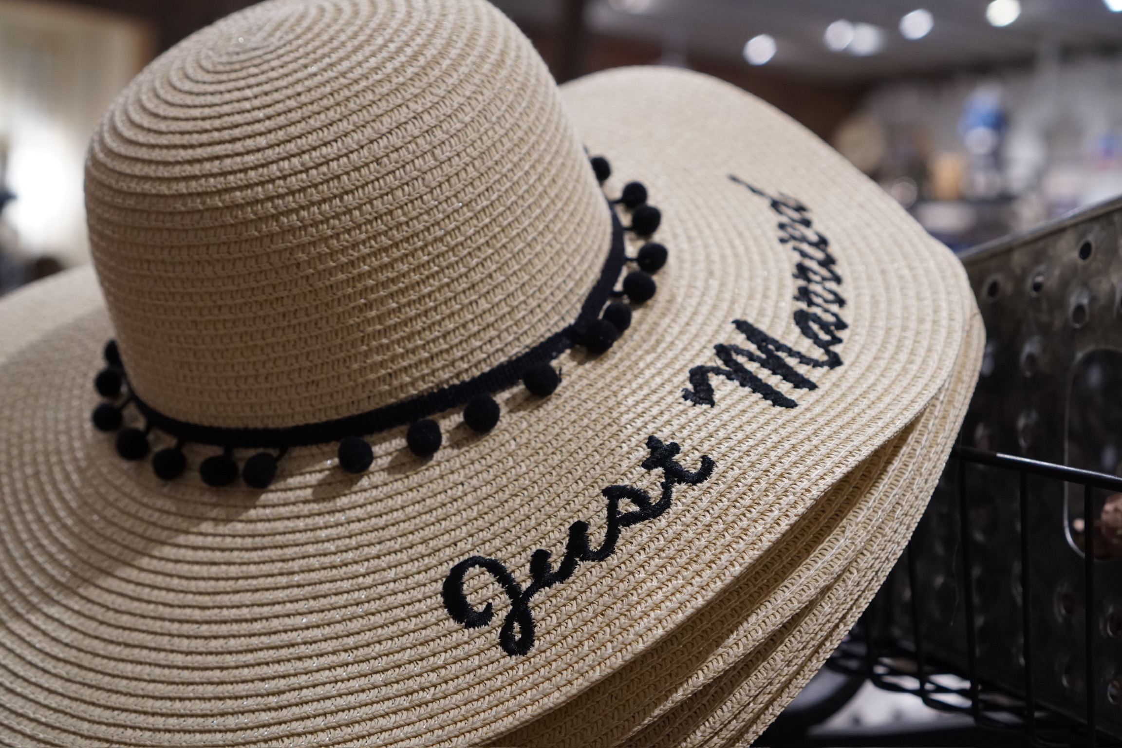 A beige sunhat with black pom-pom trim and the words "Just Married" embroidered on the brim rests on display indoors.