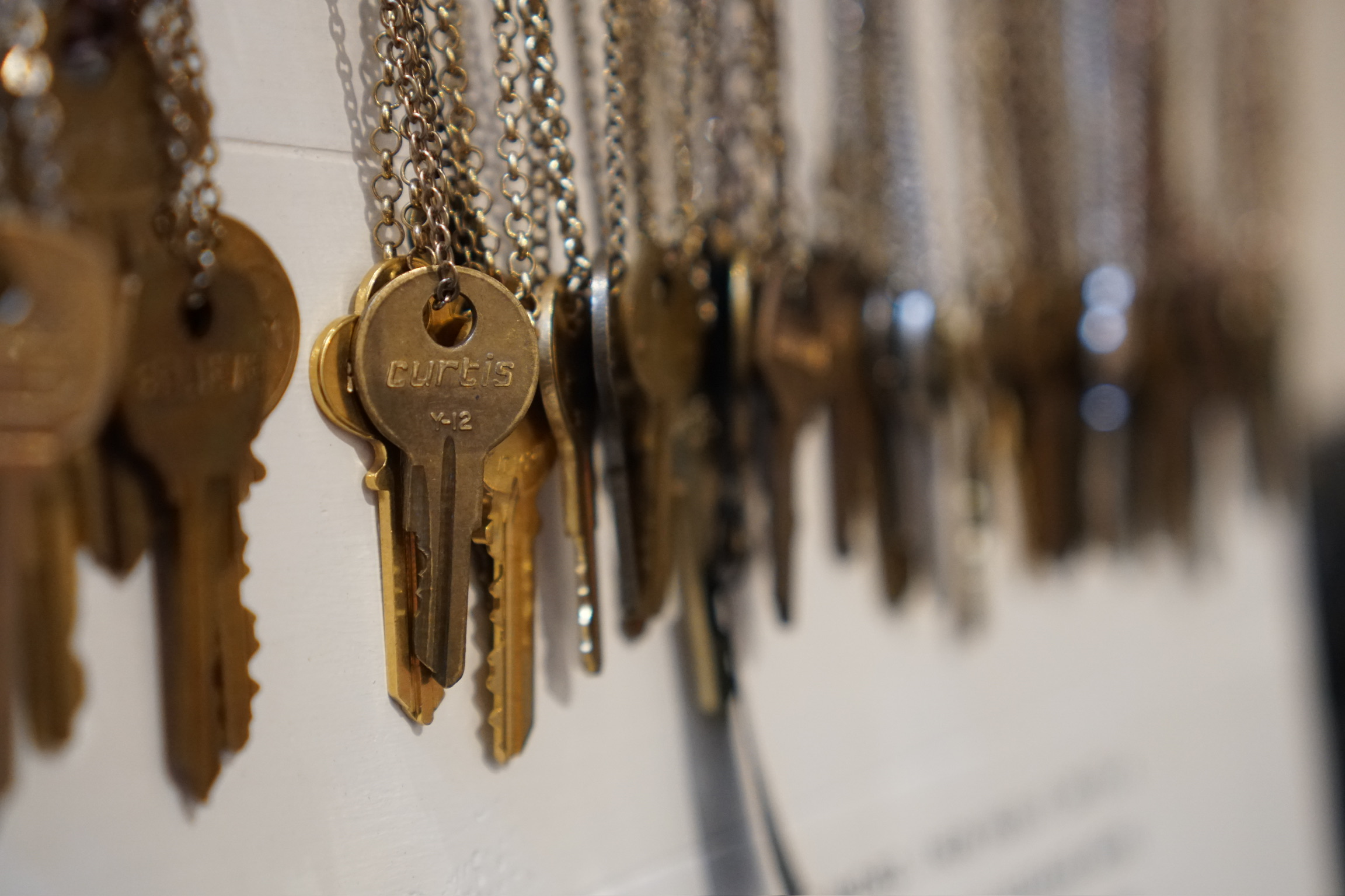 A row of brass keys hanging on metal chains against a white surface, with the word "CUTHS" visible on one key in focus.