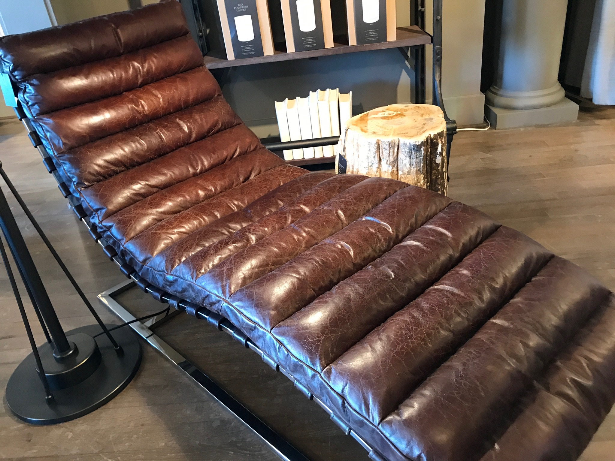 A brown leather chaise lounge with a ribbed cushion sits on a wooden floor next to a tree stump side table and a bookshelf.