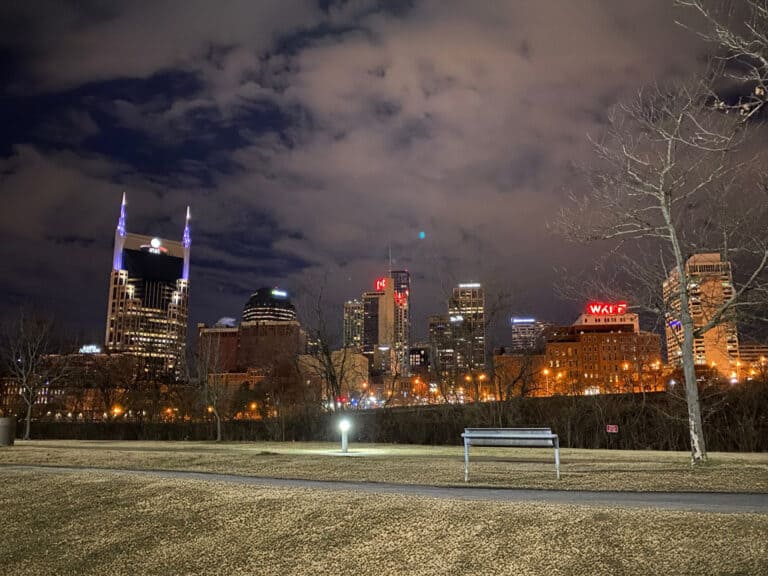 Nashville city skyline at night with illuminated buildings, cloudy sky, leafless trees, and a park bench in the foreground.