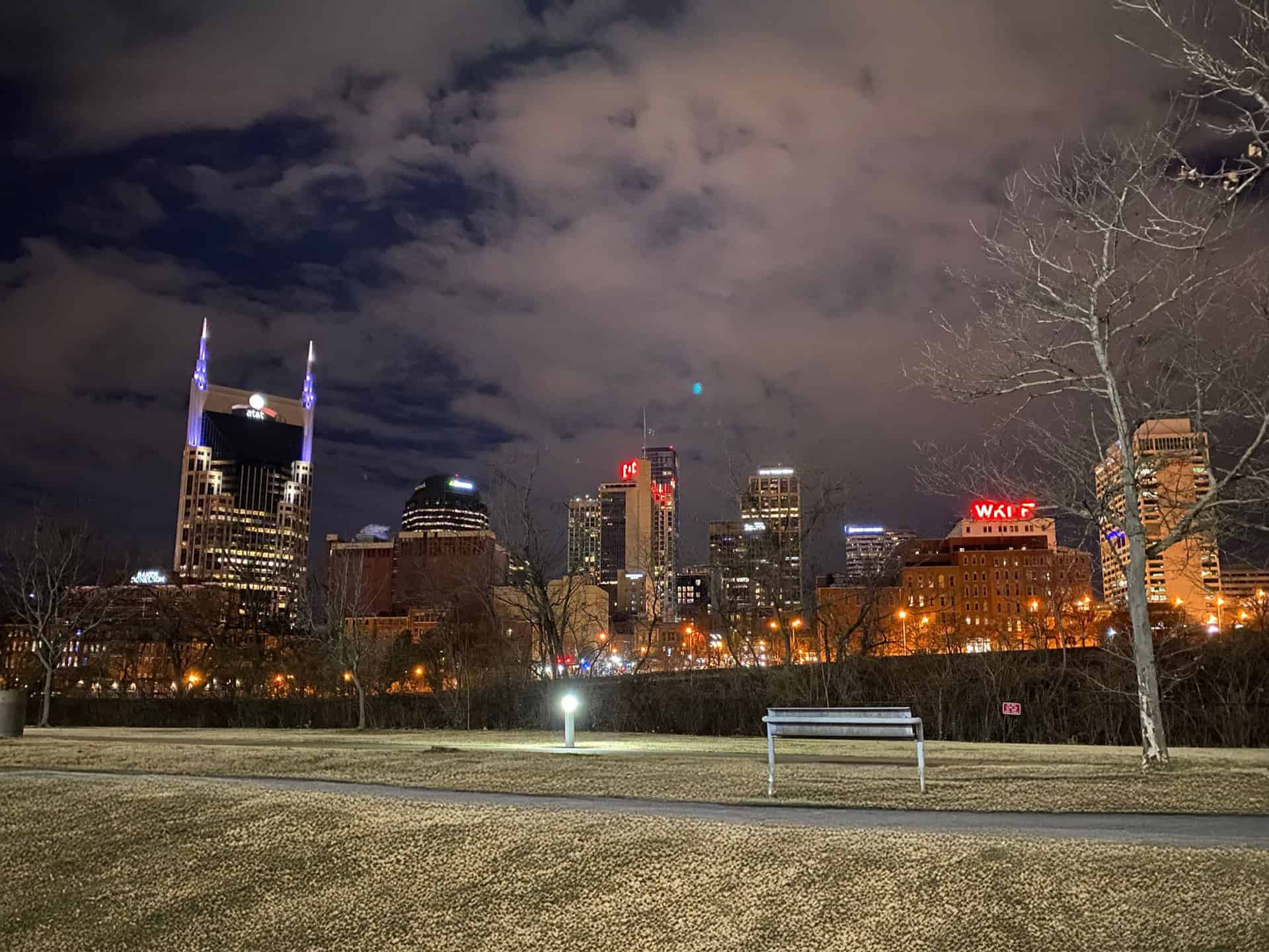 Nashville city skyline at night with illuminated buildings, cloudy sky, leafless trees, and a park bench in the foreground.