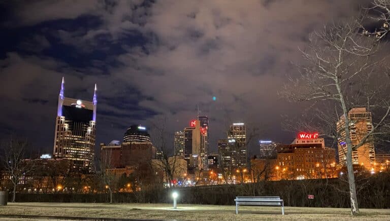 Nashville city skyline at night with illuminated buildings, cloudy sky, leafless trees, and a park bench in the foreground.