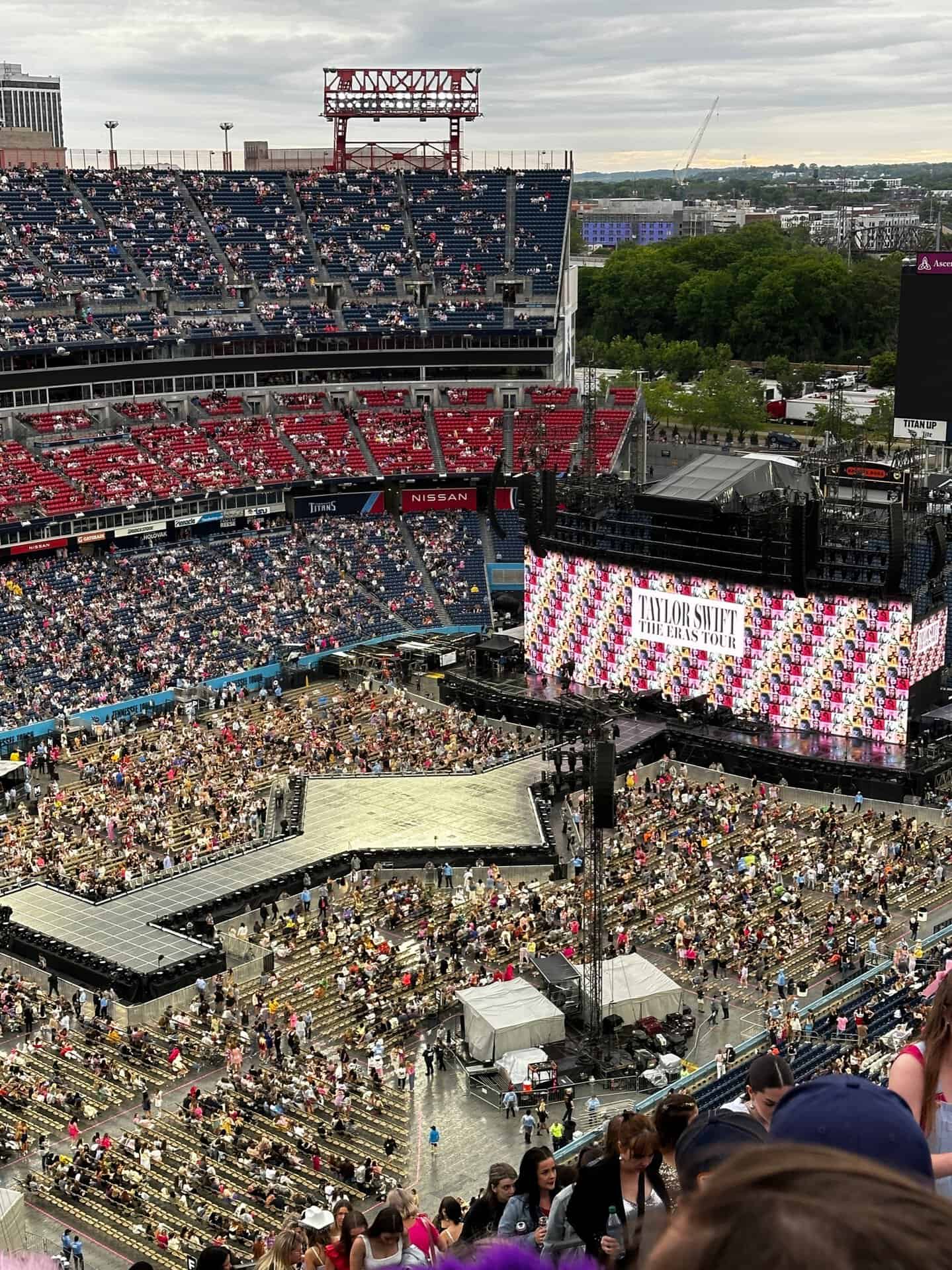 A stadium in Nashville buzzes with a large crowd, all eyes on the stage displaying "Taylor Swift The Eras Tour," ready for an unforgettable concert experience.