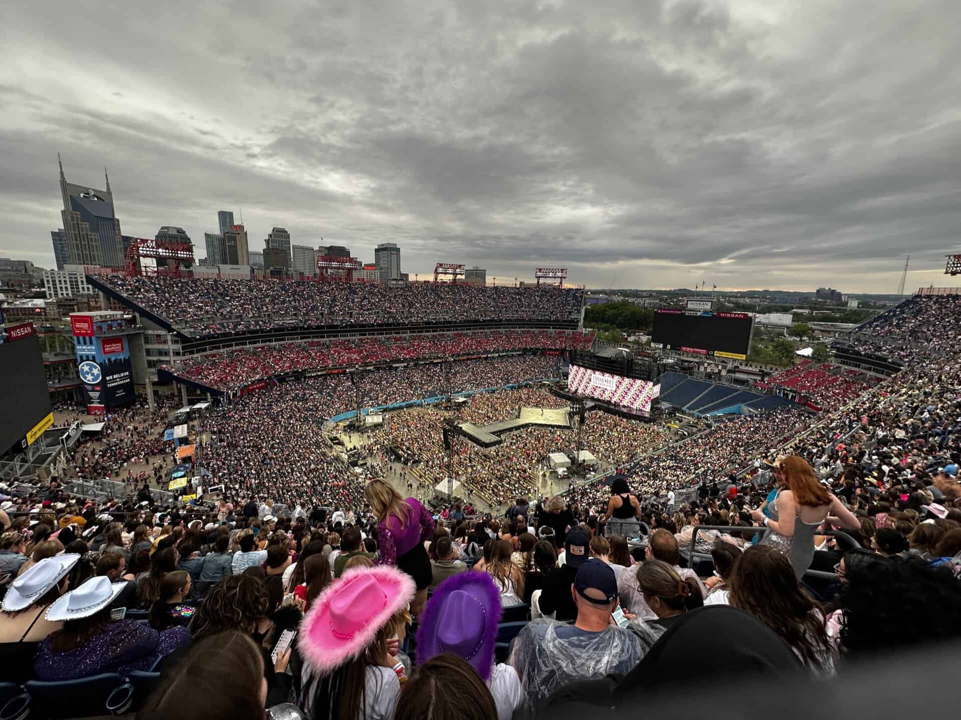 A large crowd fills an outdoor stadium in Nashville for a concert, with the city skyline visible in the background under a cloudy sky. People are seated and standing, some wearing hats.