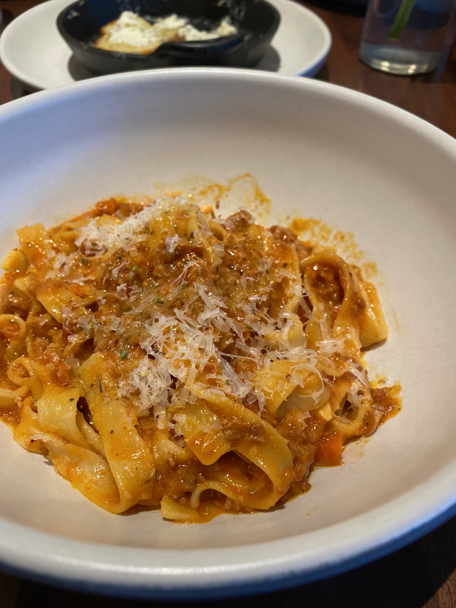 A bowl of tagliatelle pasta with meat sauce, topped with grated cheese, is served on a table with another dish in the background.