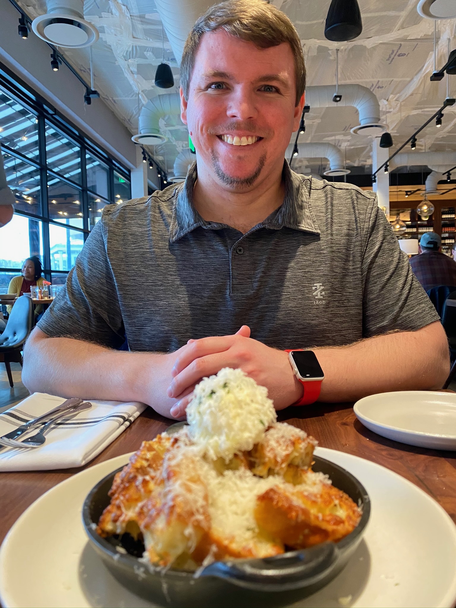 A man sits at a restaurant table smiling, with a skillet of baked pasta topped with cheese in front of him.