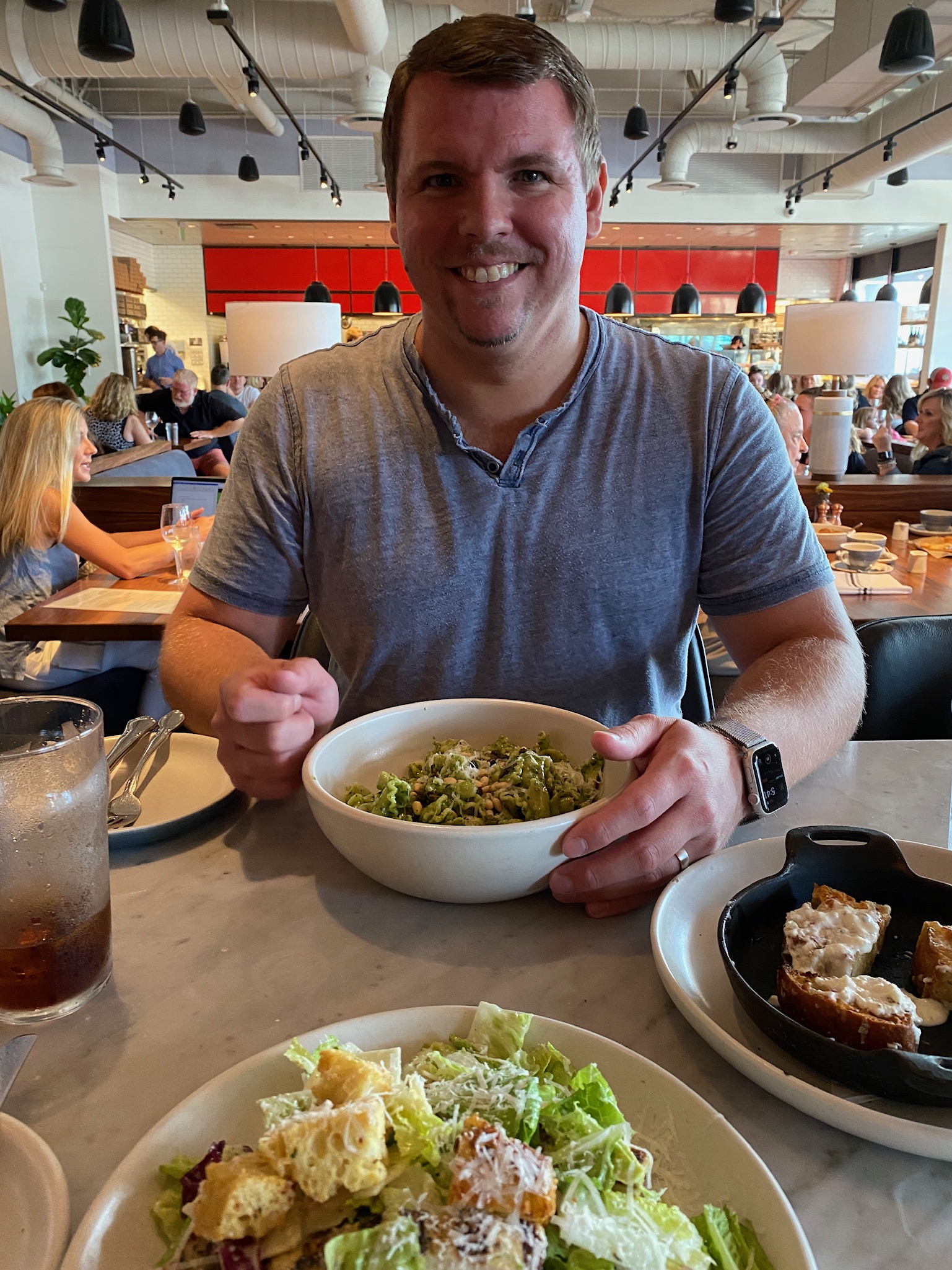 A man sits at a restaurant table smiling, holding a bowl of salad. There is another salad and a plate of bread on the table, with people dining in the background.