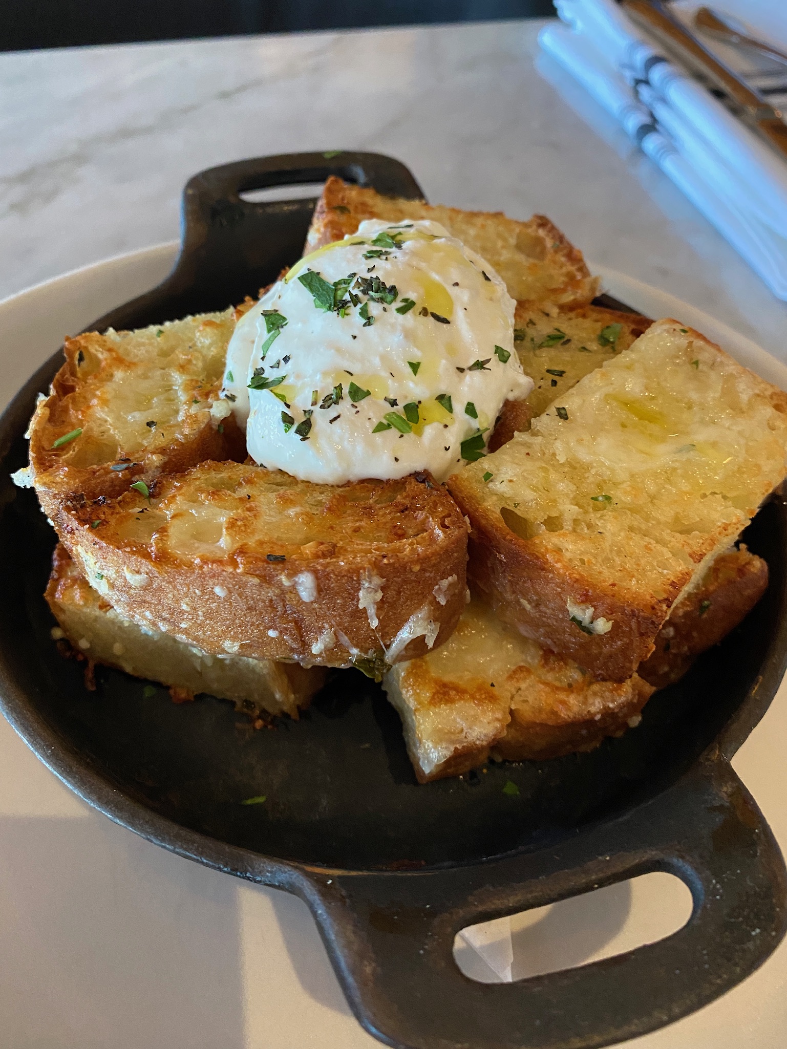 Slices of toasted bread topped with a dollop of creamy spread and sprinkled with herbs, served in a small black pan on a white plate.