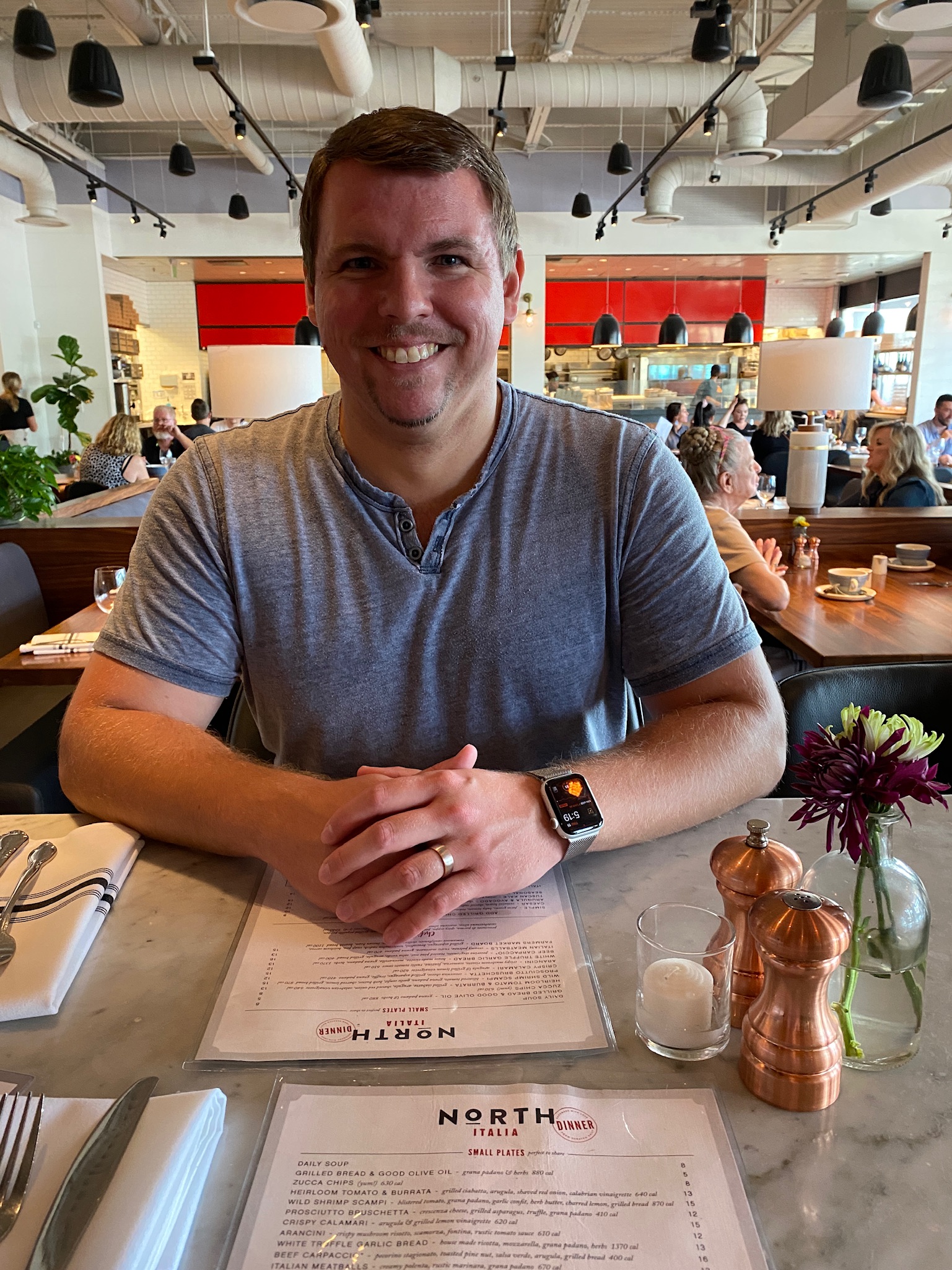 A man sits at a restaurant table with menus, cutlery, flowers, and condiments, smiling at the camera. Other diners are visible in the background.