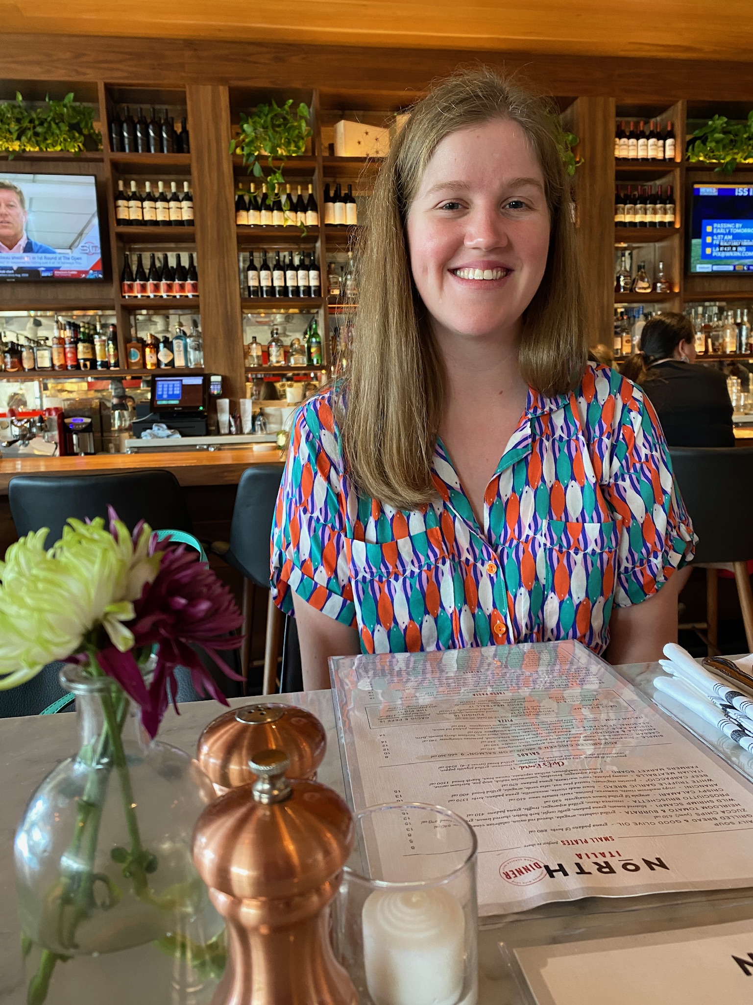 A woman with long hair sits at a restaurant table with a menu, salt and pepper shakers, a candle, and flowers; shelves of bottles and TVs are visible in the background.