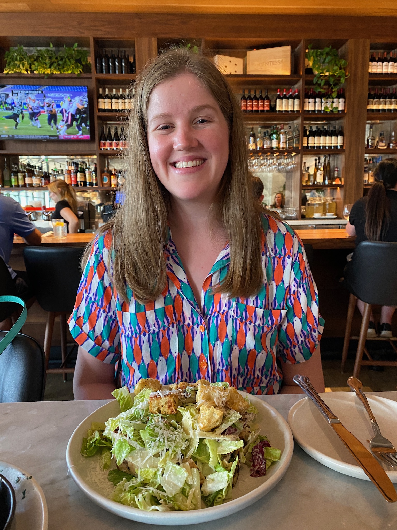 A woman sits at a restaurant table, smiling, with a plate of salad in front of her; shelves of wine bottles and a TV are visible in the background.