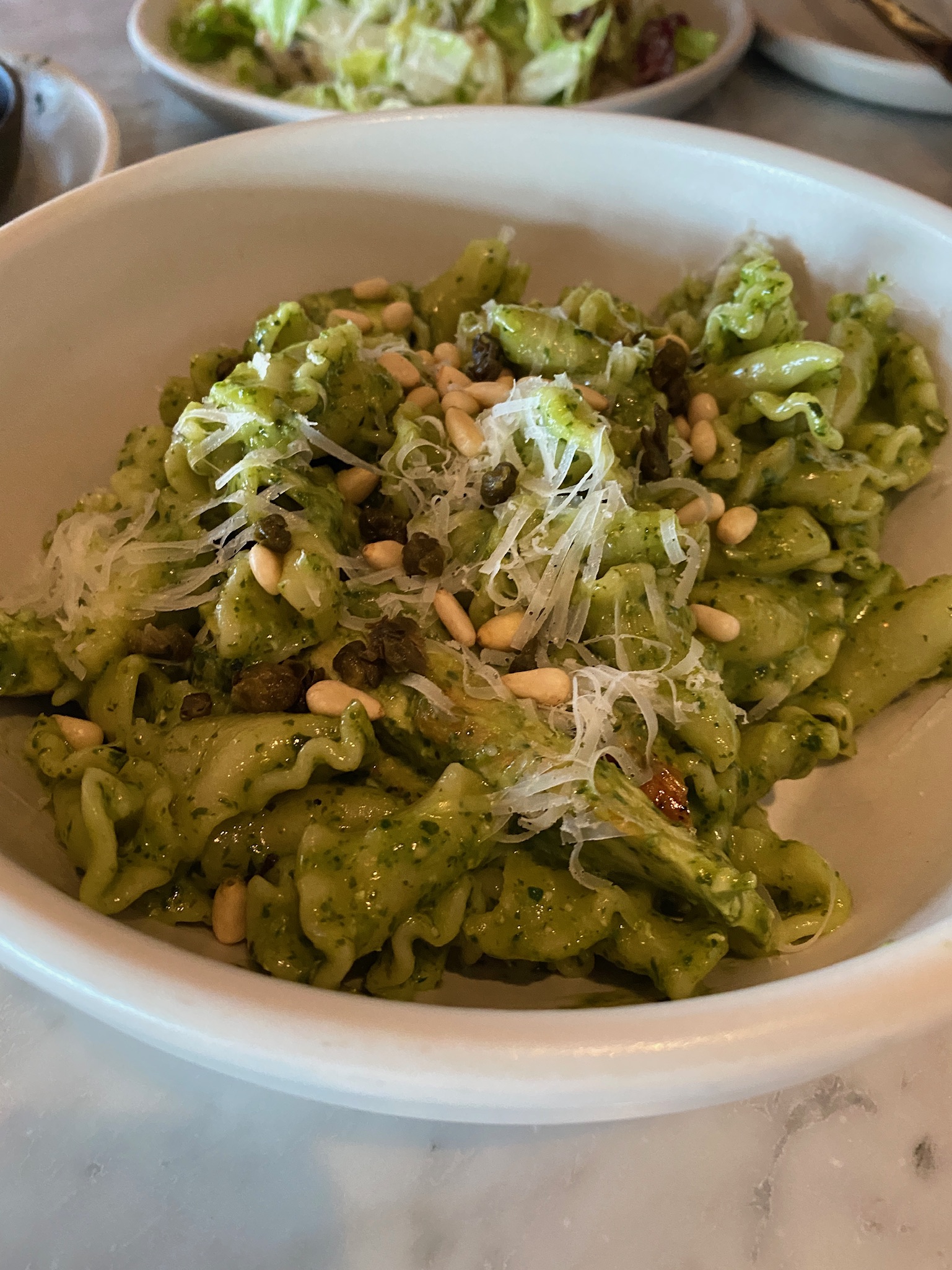 A bowl of pasta with green pesto sauce, grated cheese, pine nuts, and capers, served on a white plate.