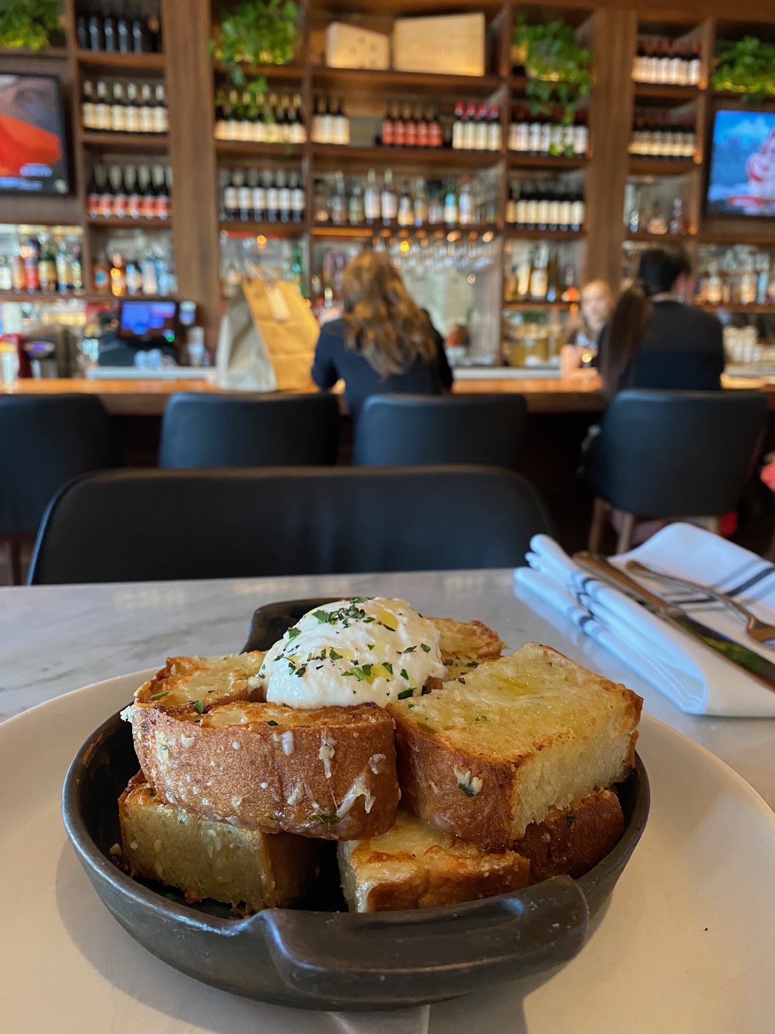 A plate of sliced toasted bread topped with a dollop of herbed butter sits on a restaurant table; bar and shelves with bottles in the background.