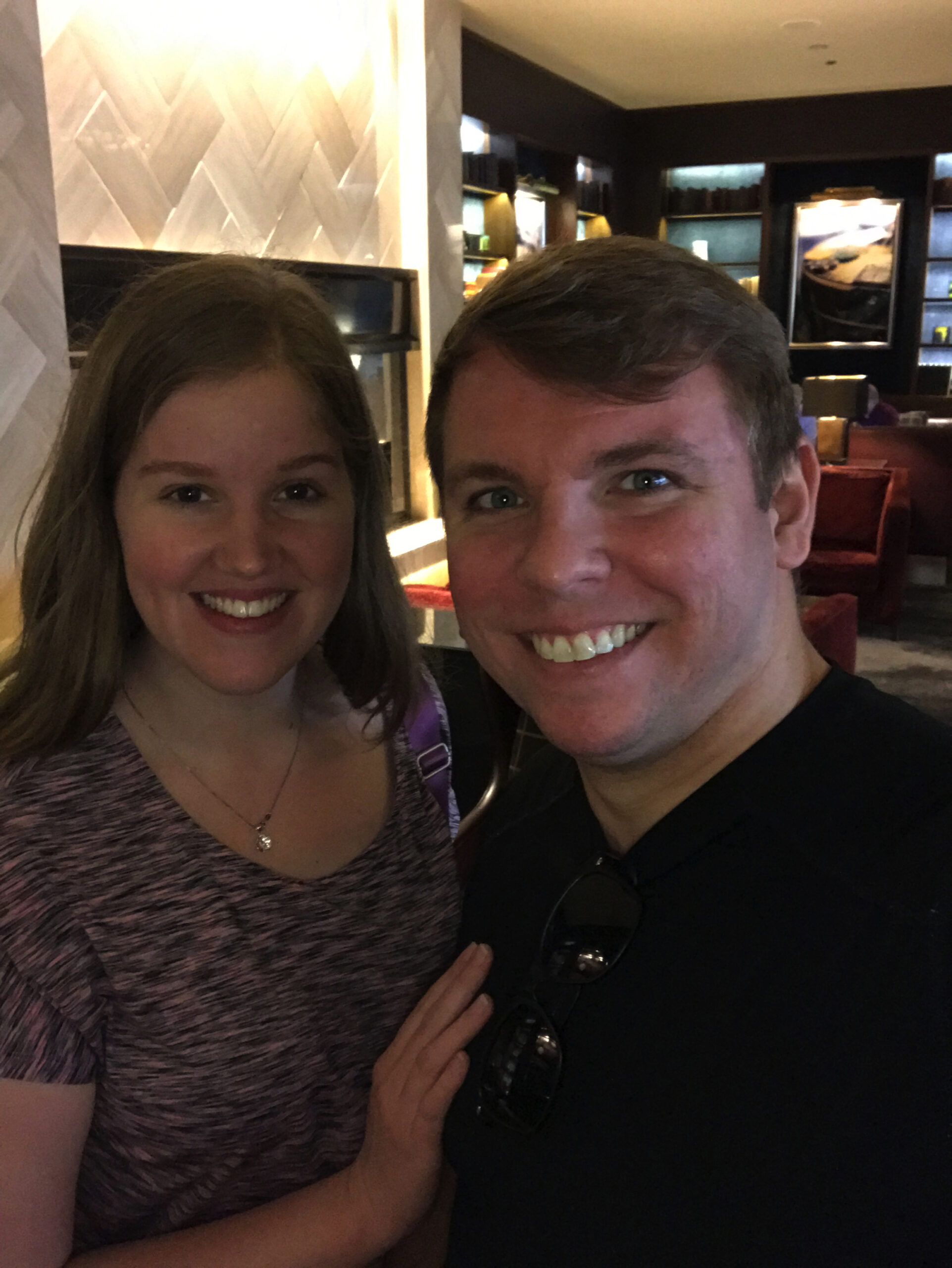 A woman and a man smile at the camera indoors, standing close together in a warmly lit room with shelves and seating in the background.