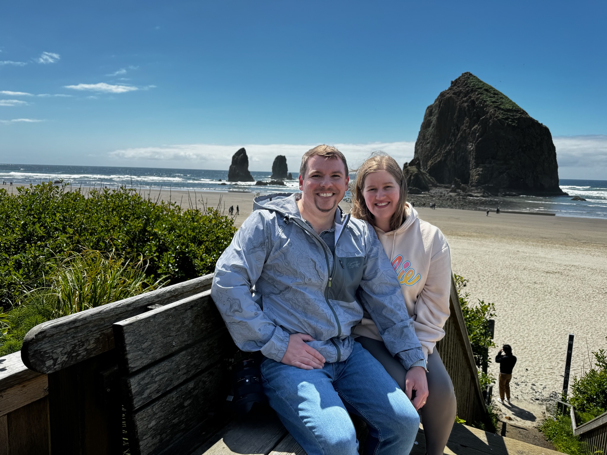 A man and woman sit on a wooden bench at a sandy beach with large rock formations and the ocean in the background under a partly cloudy sky.