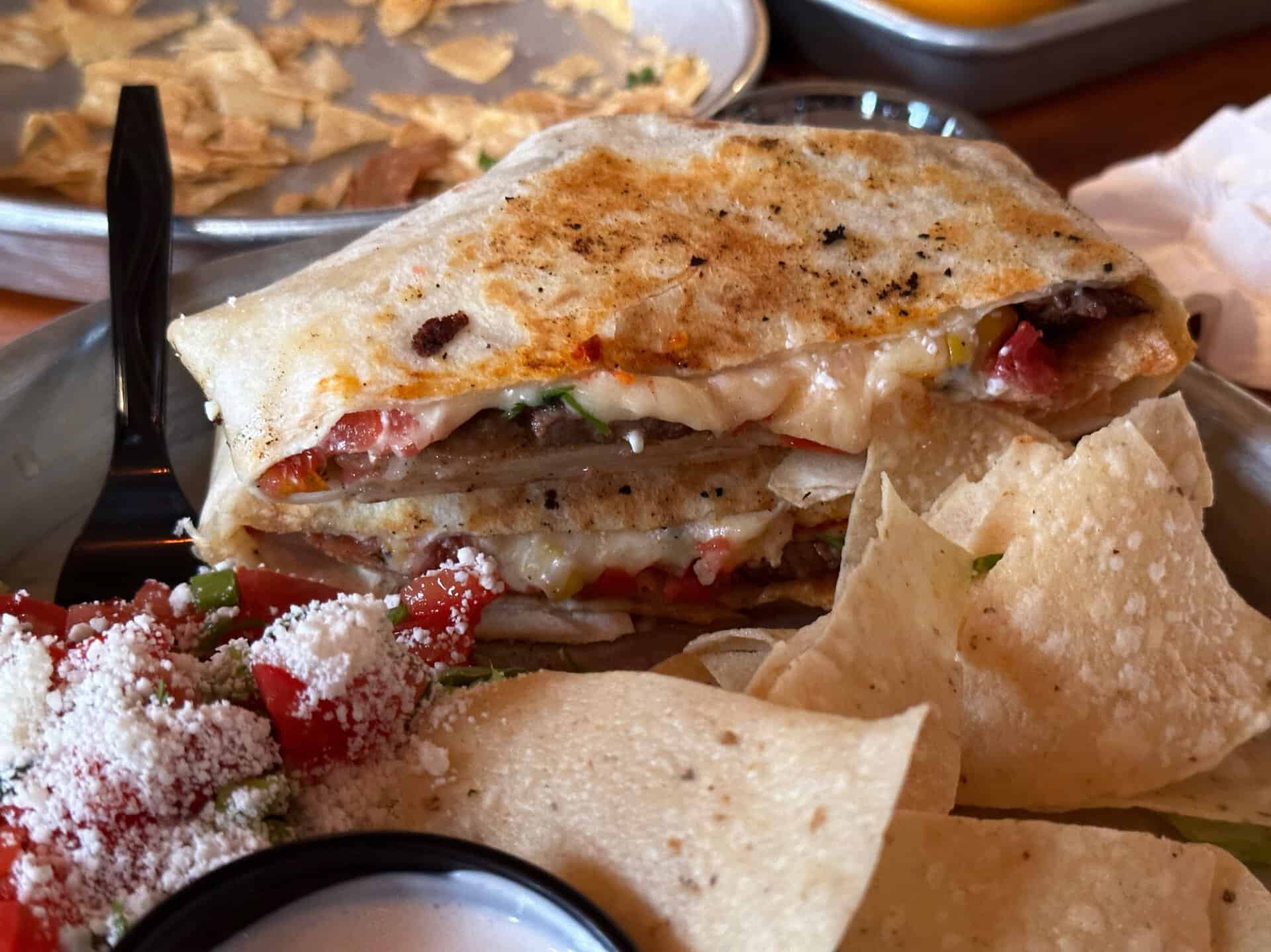 A Nashville quesadilla stacked with melted cheese and vegetables, served on a metal plate with tortilla chips and a side of salsa.