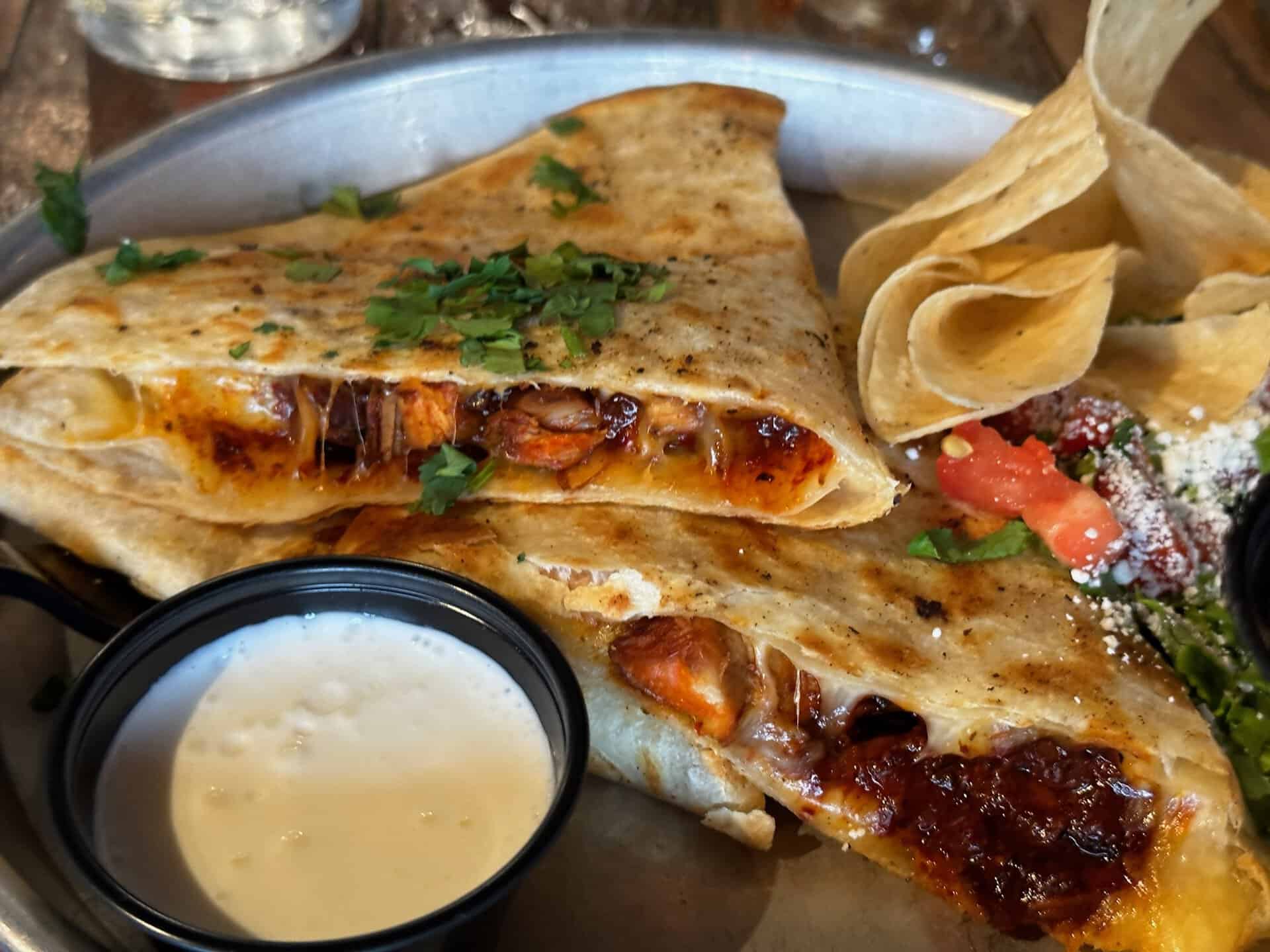 A Nashville-inspired quesadilla with melted cheese and grilled chicken, garnished with cilantro. Served alongside Southern-style tortilla chips, a side of creamy white dipping sauce, and a small salad topped with tomatoes and cheese.