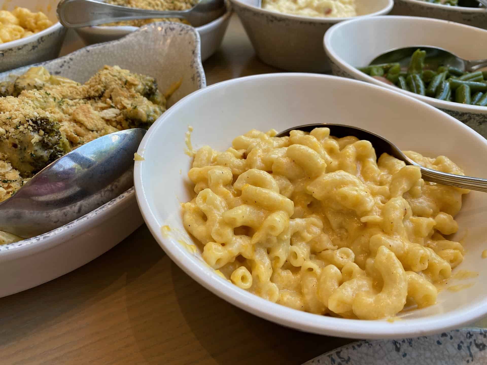 A bowl of macaroni and cheese with a spoon, surrounded by dishes of vegetables and other foods on a wooden table.