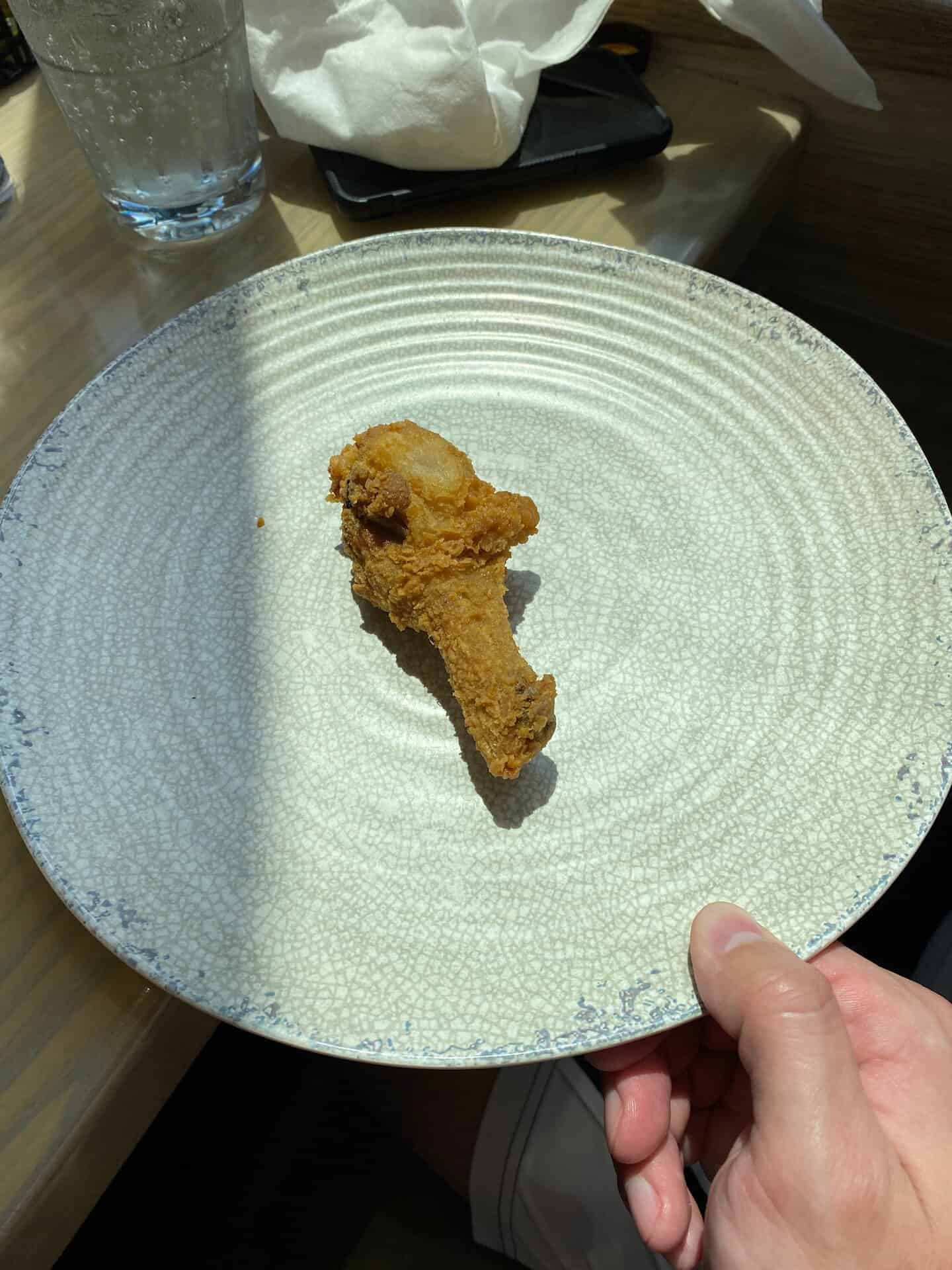 A single piece of fried chicken on a textured round white plate, held by a hand above a wooden table with a glass and napkin in the background.