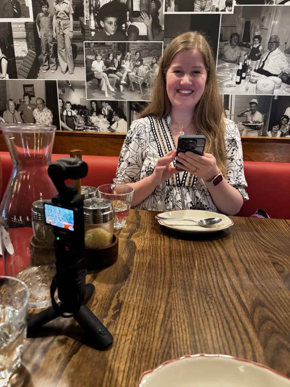 A woman sits at a Nashville restaurant table, smiling as she holds her phone. A small camera on a tripod rests beside her, capturing the moment. Black and white photos adorn the wall behind her, adding a touch of charm to the scene.