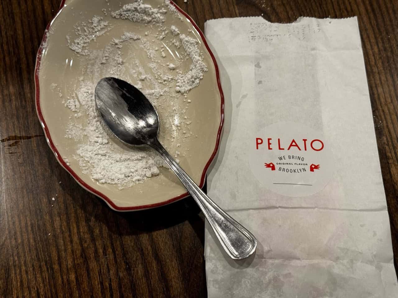 An empty dessert plate with powdered sugar remnants and a spoon rests next to a white paper bag labeled "Pelato" on a wooden table in a cozy Nashville café.