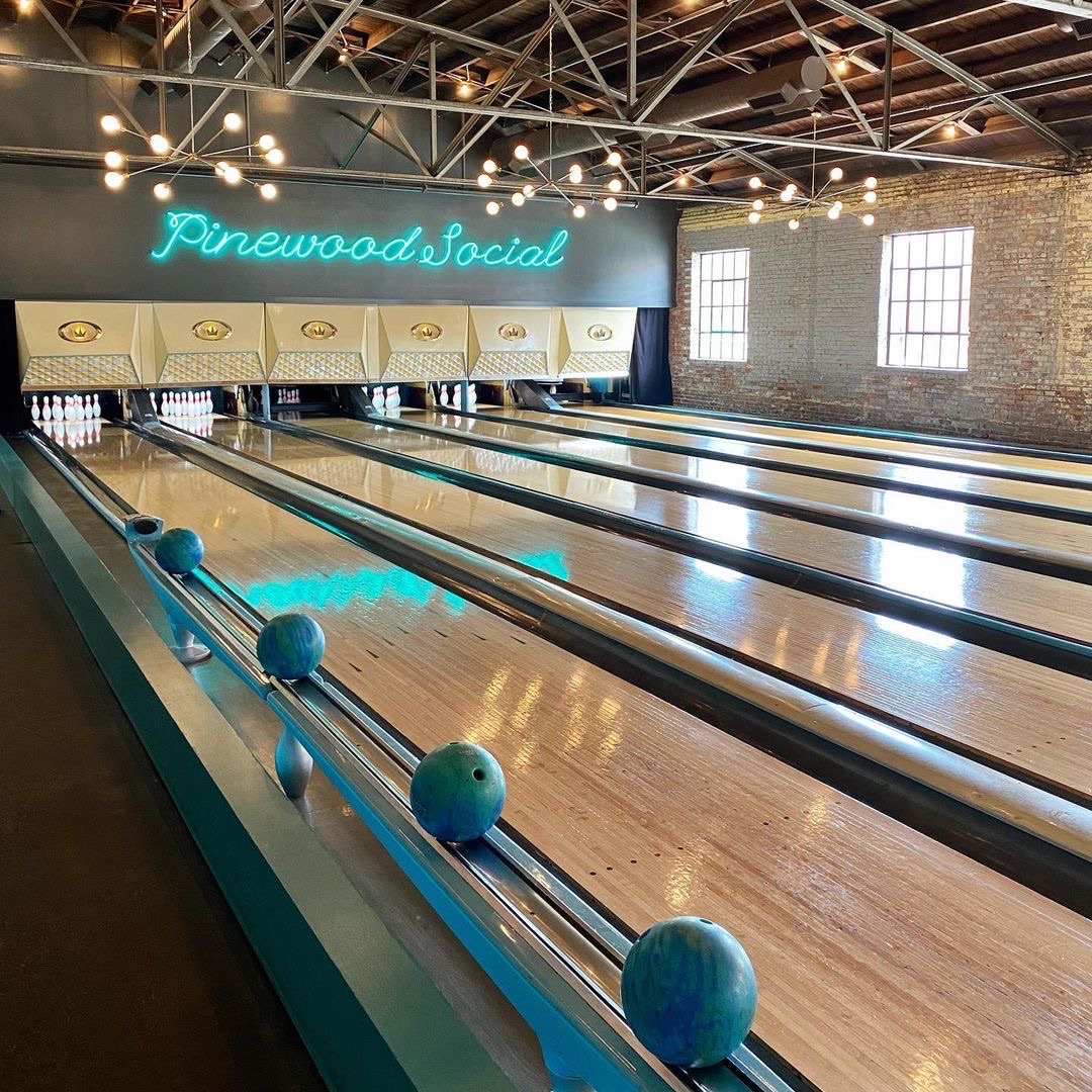 Four lane vintage-style bowling alley with blue bowling balls, wooden floors, exposed brick walls, and a neon "Pinewood Social" sign above the lanes.