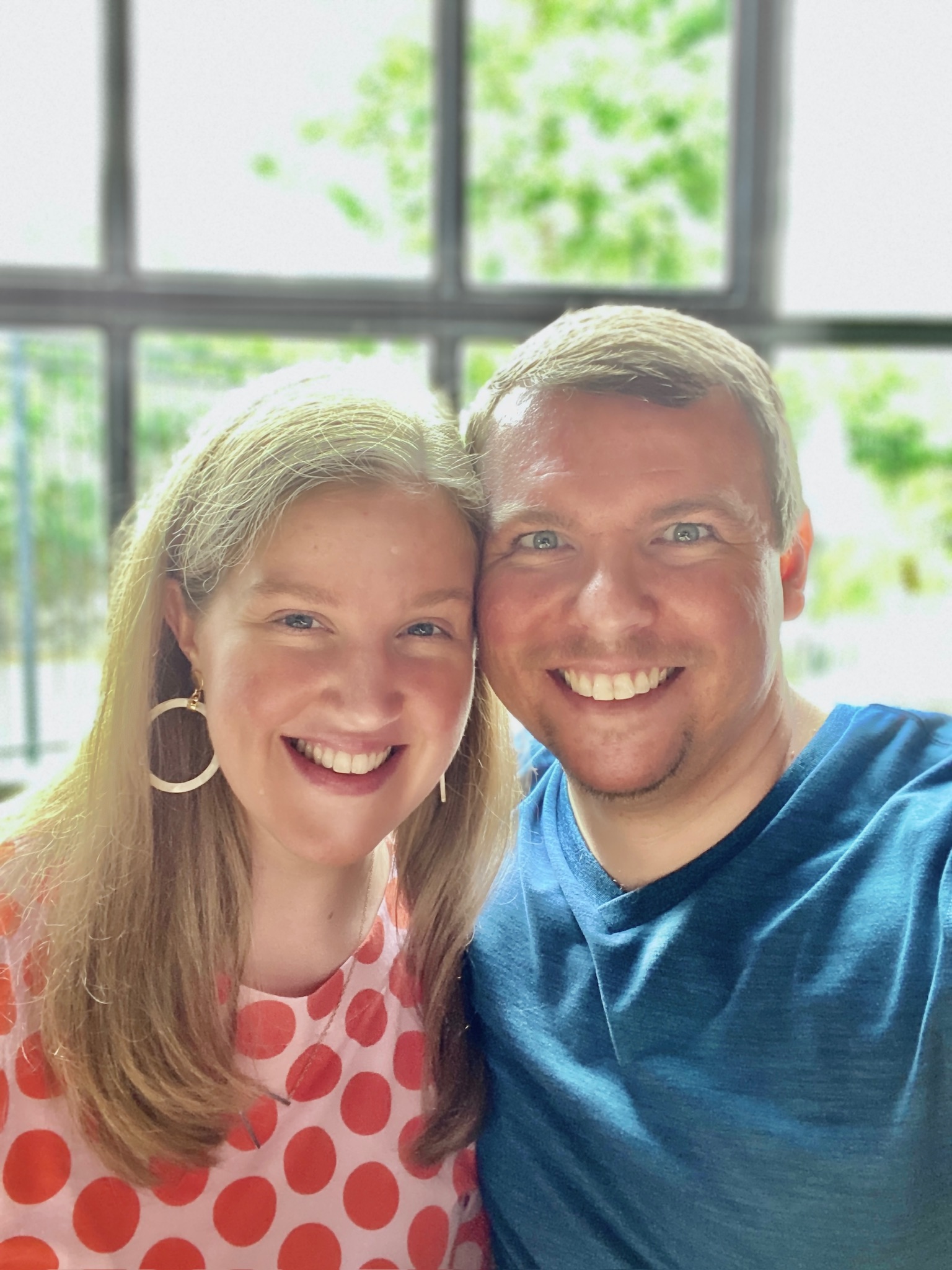 A woman in a red polka dot top and a man in a blue shirt smile together for a selfie indoors with a bright window in the background.