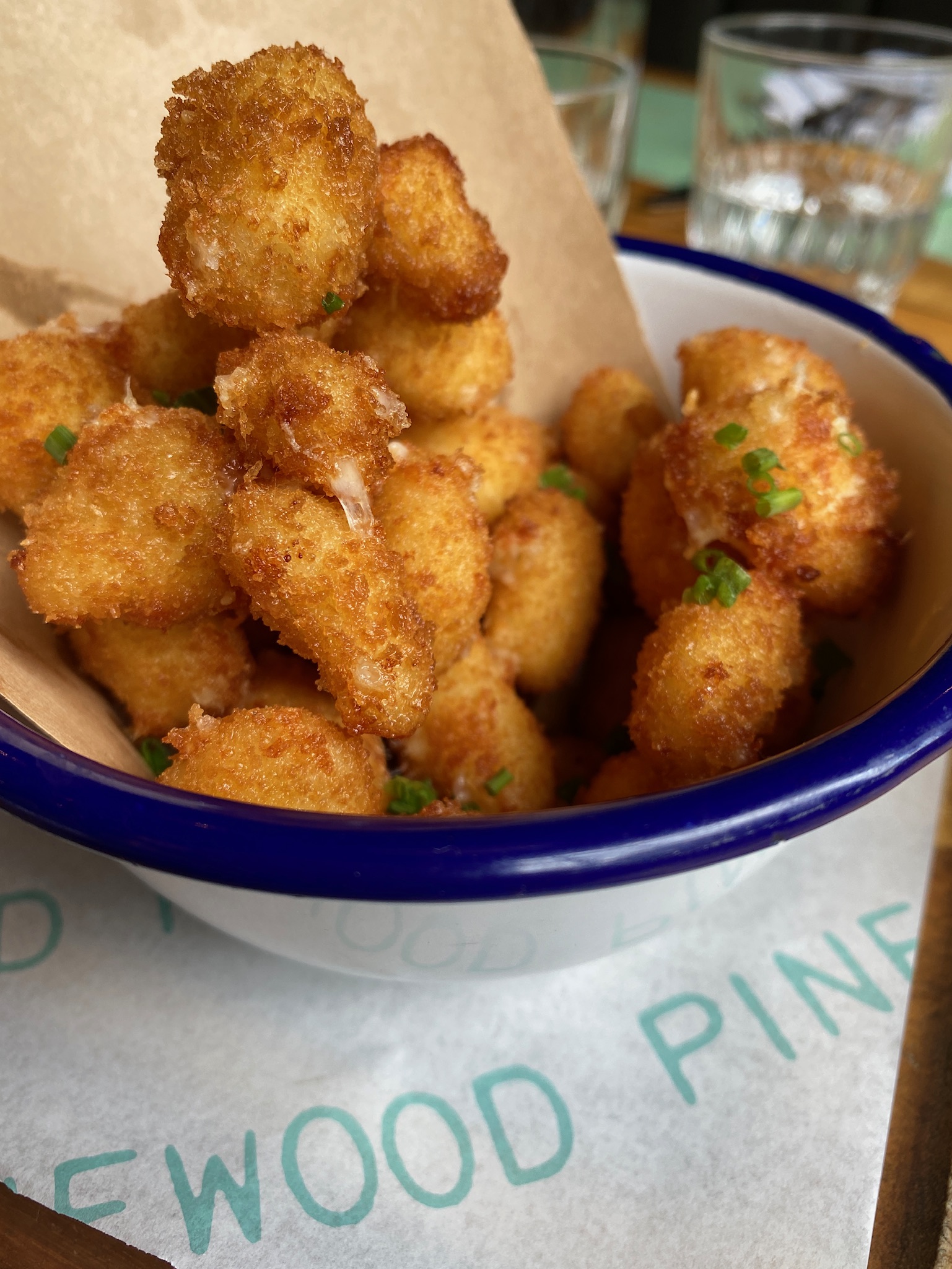 A bowl filled with crispy, golden-brown breaded cheese curds, garnished with chopped chives, sits on a table next to drinking glasses.