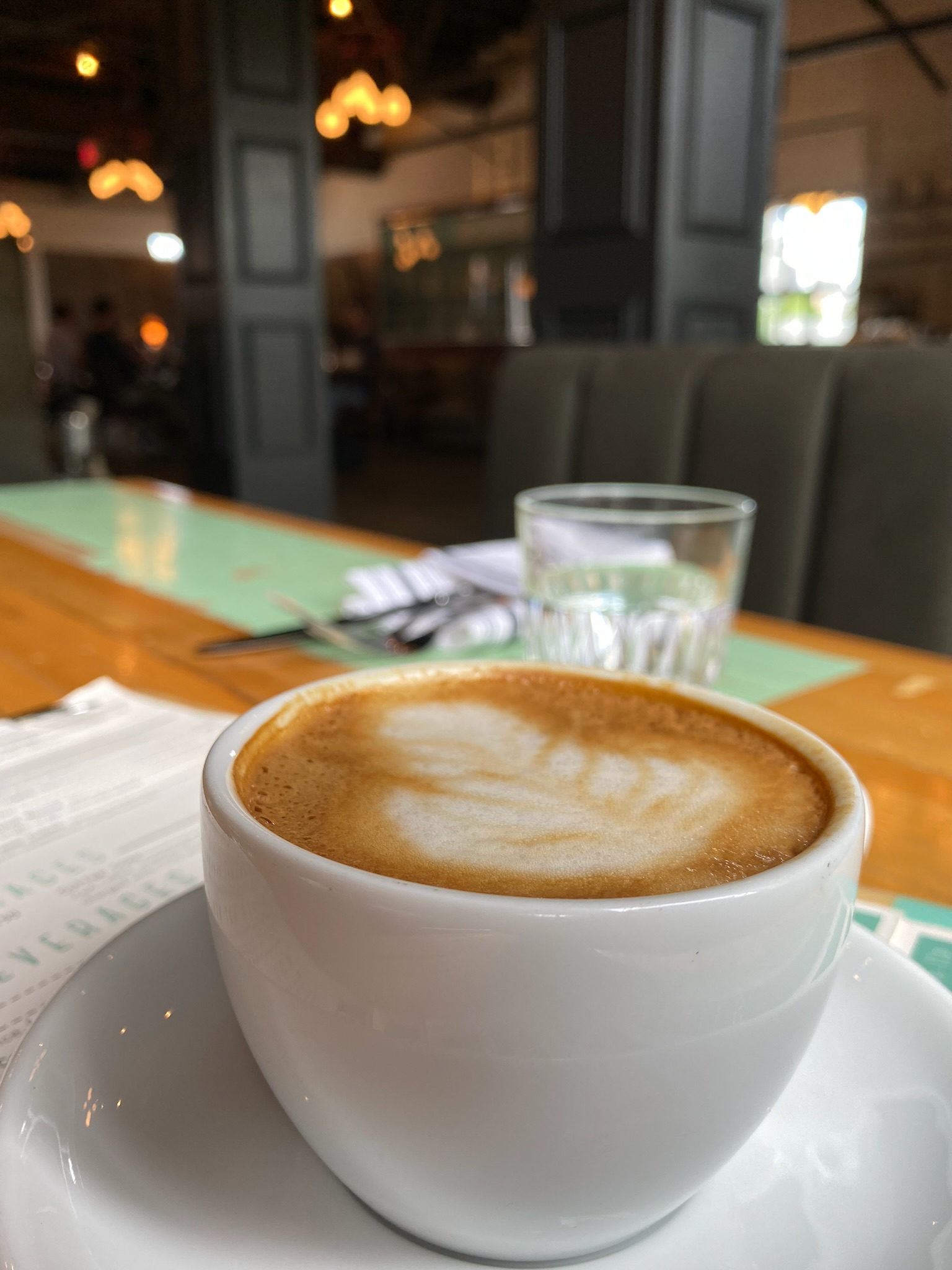 A cup of cappuccino sits on a wooden table next to a glass of water, with a menu and cutlery in the background at a restaurant.