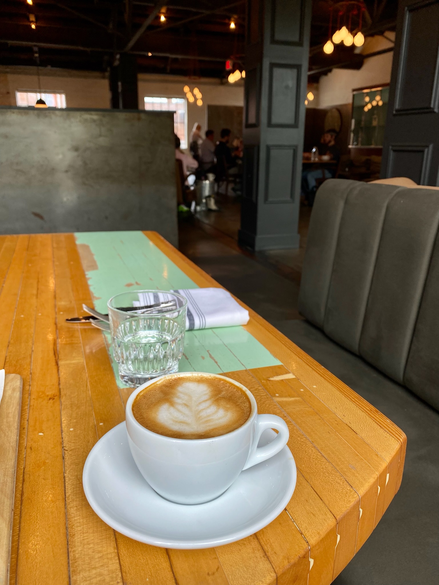 A cup of latte with latte art, a glass of water, and a folded napkin on a wooden table in a modern café with people seated in the background.