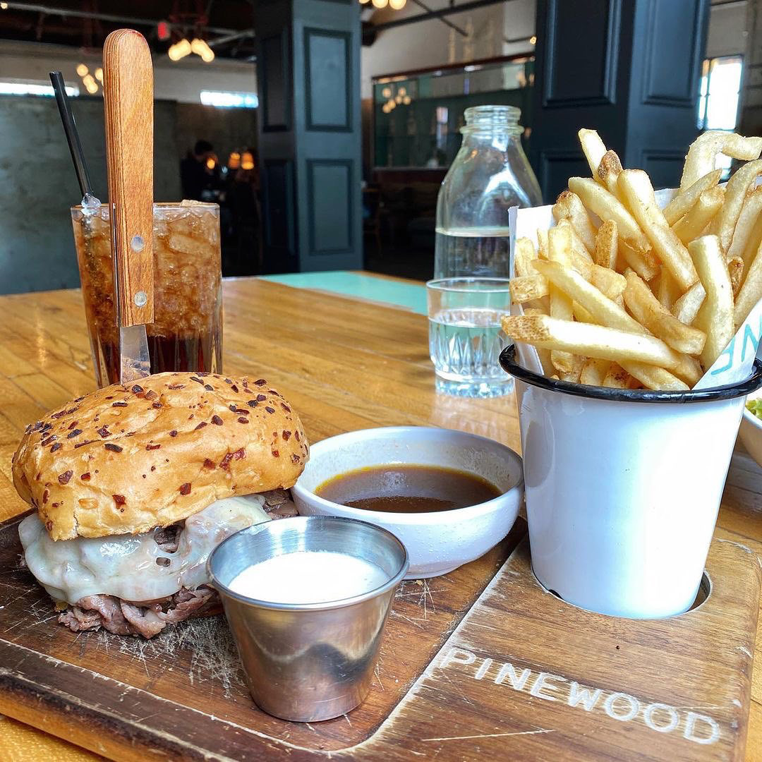 A sandwich with melted cheese, a cup of sauce, and a side of fries are served on a wooden board with a drink and a bottle of water on the table.