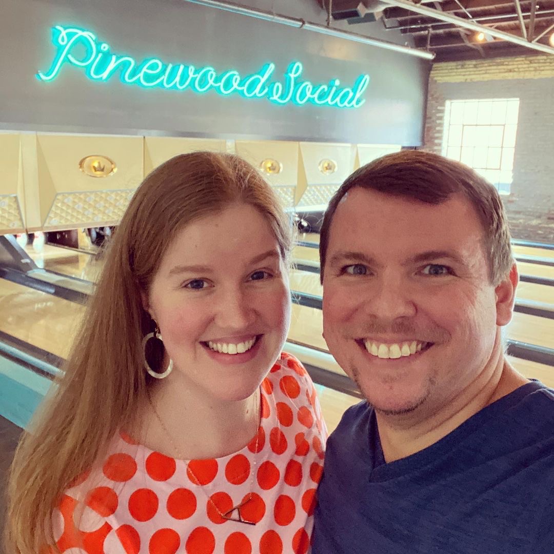 Two people smiling in front of bowling lanes at Pinewood Social, with a neon sign for the venue visible in the background.