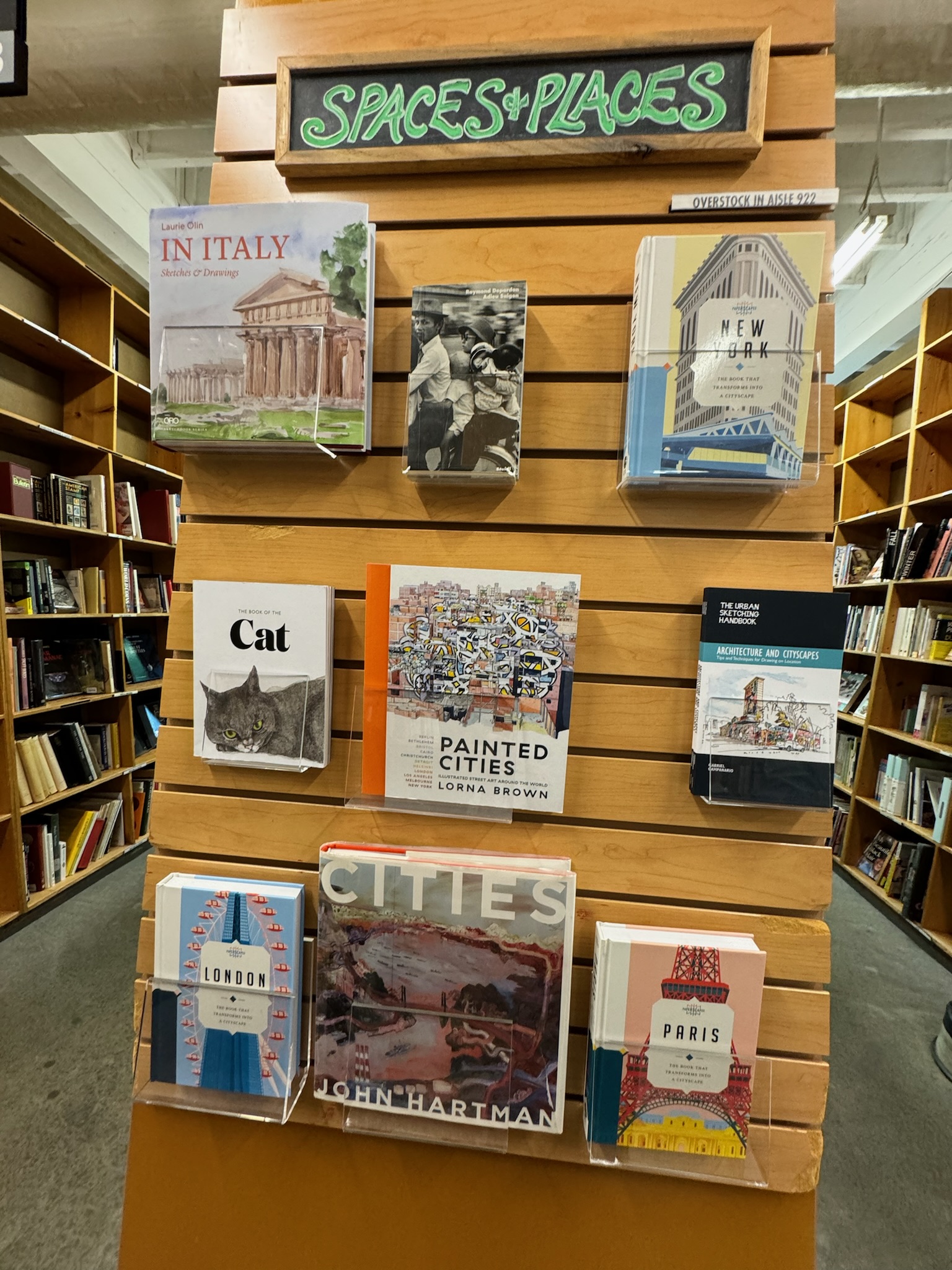 A bookshelf display labeled "Spaces + Places" featuring books about various cities and locations, including Italy, New York, London, Paris, and others, in a bookstore setting.
