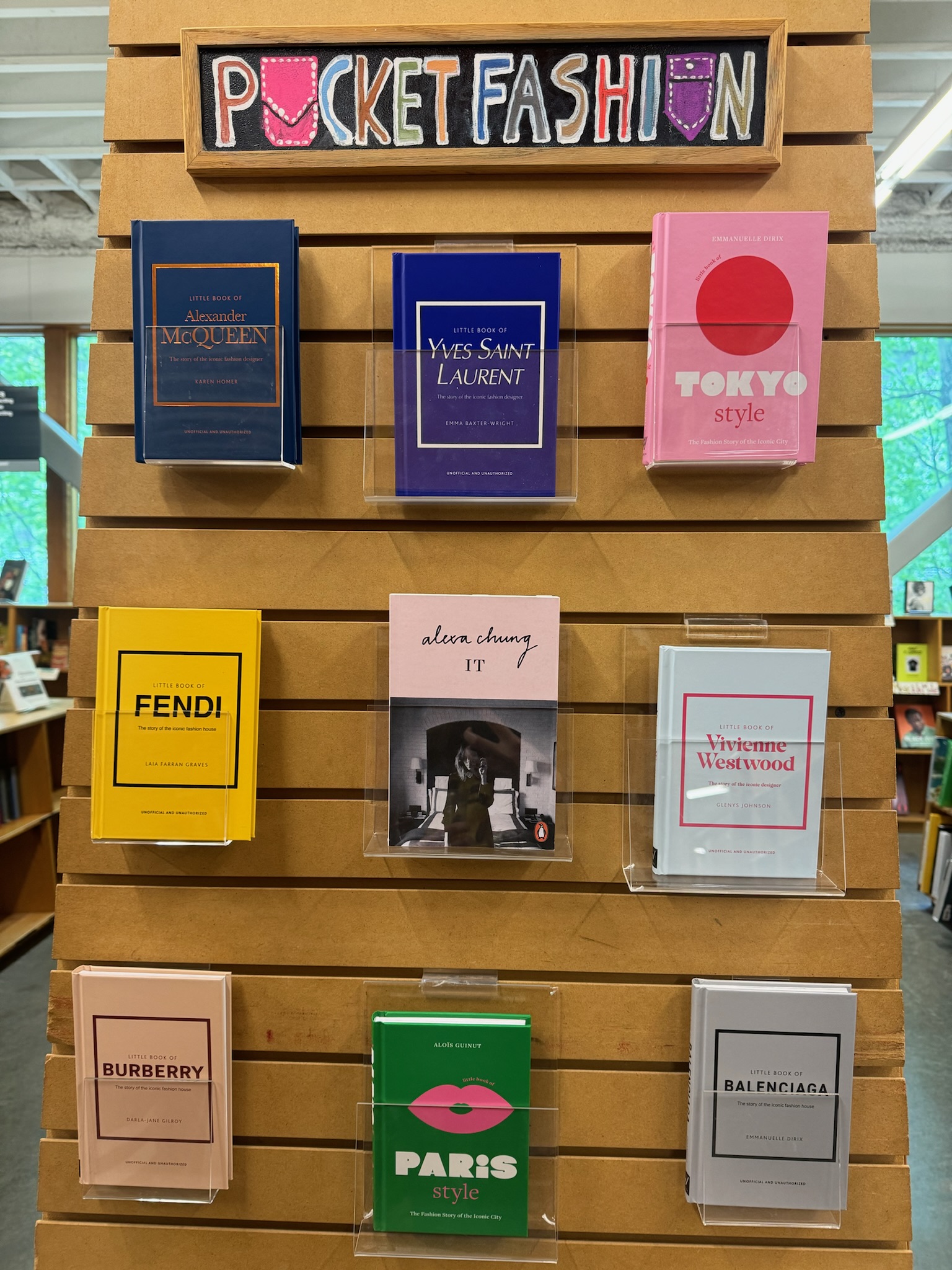 A bookstore display features nine fashion books arranged in a grid beneath a colorful "Pocket Fashion" sign on a wooden panel.