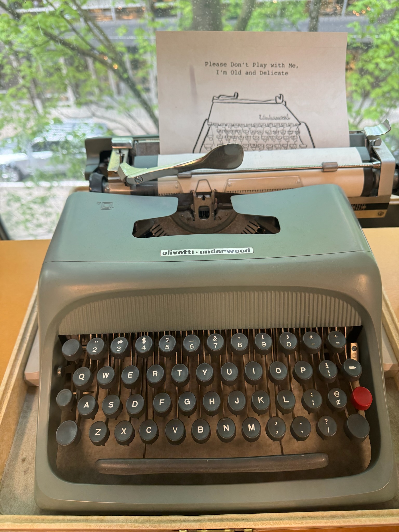 A gray Olivetti Underwood typewriter sits on a desk with a note reading, "Please Don't Play with Me, I'm Old and Delicate," placed in the paper holder.