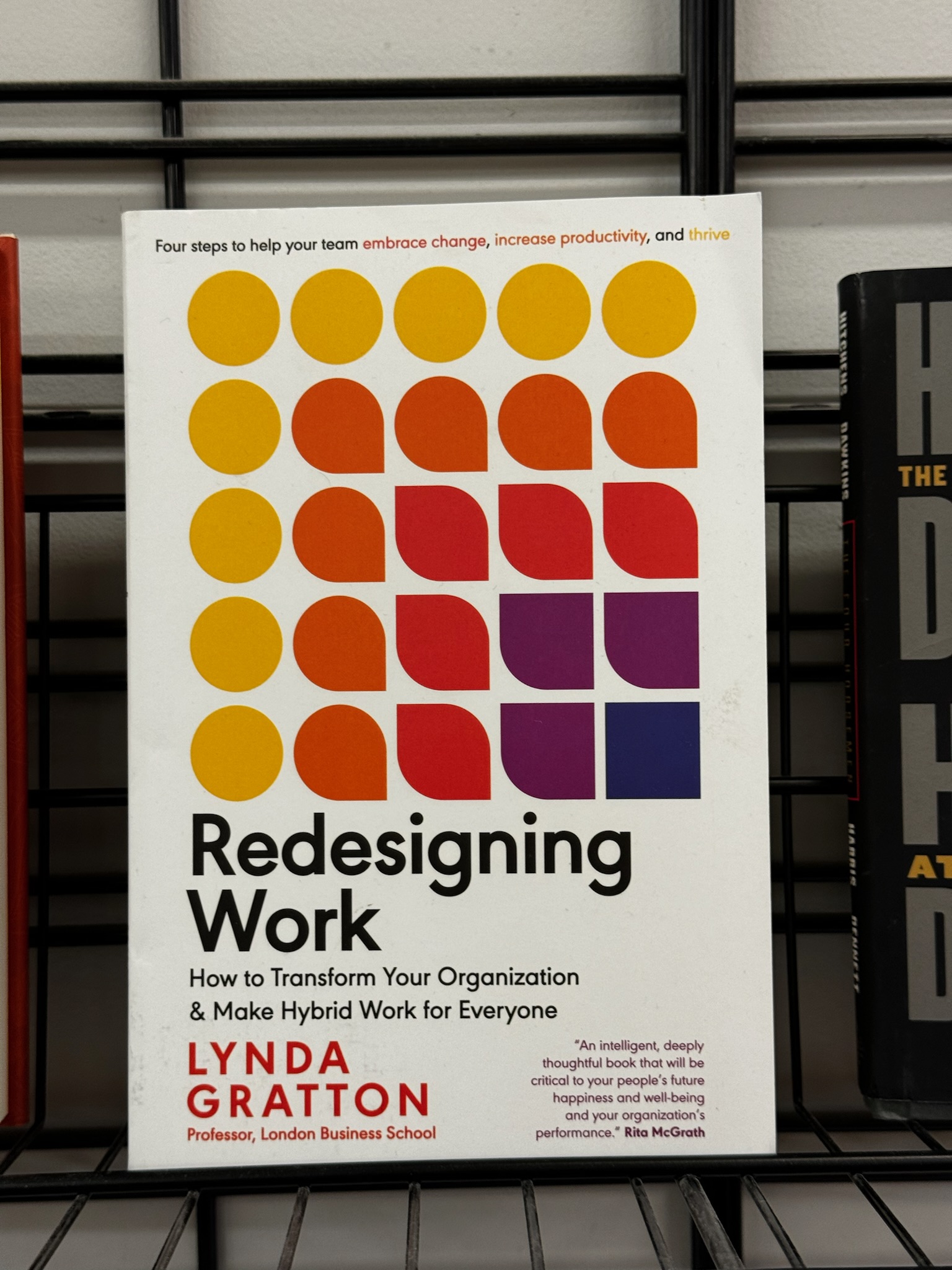 The book "Redesigning Work" by Lynda Gratton is displayed upright on a black wire shelf, showing a white cover with orange and purple geometric designs.