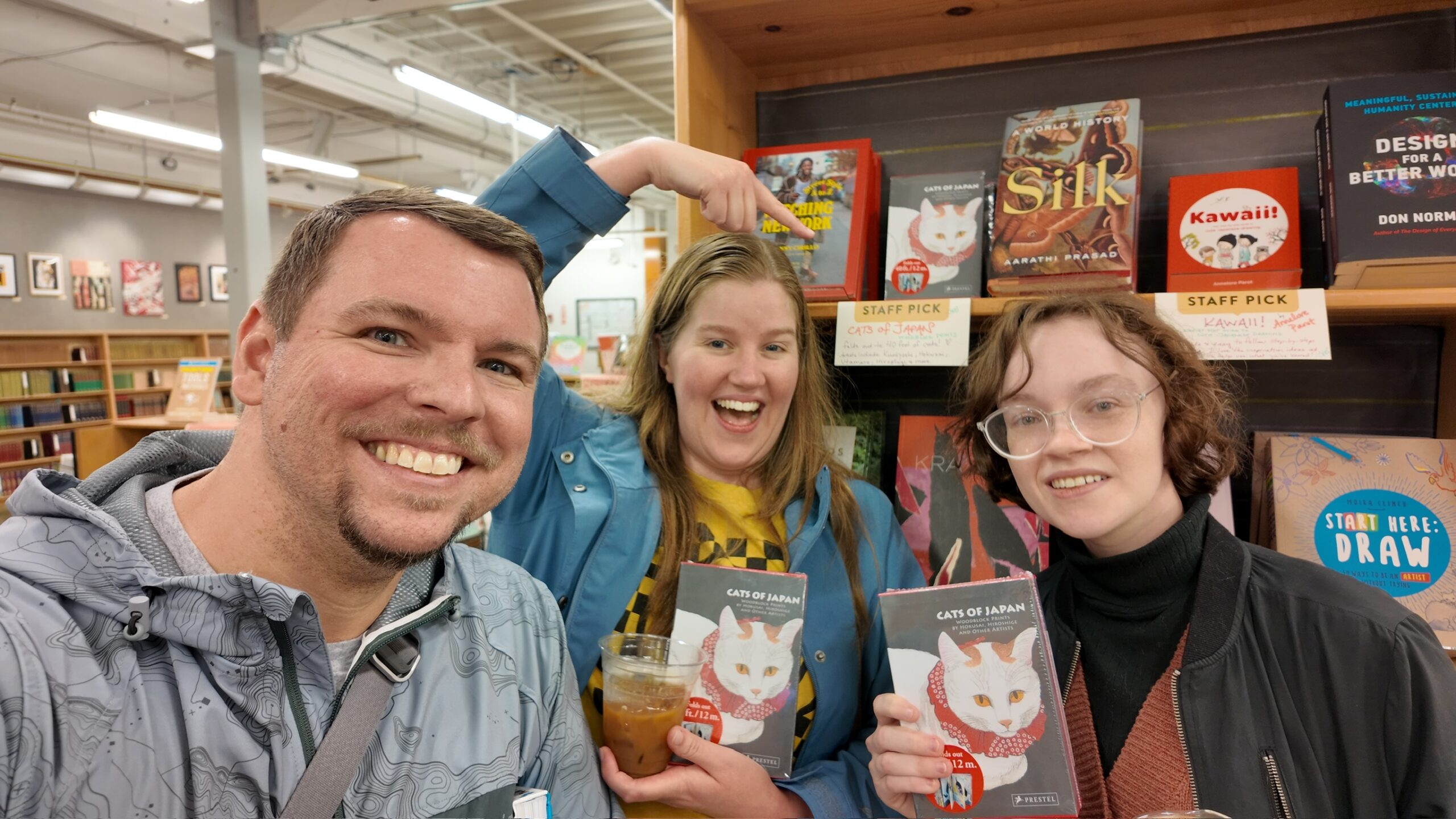 Three people smile in a bookstore, holding copies of “Cats of Japan.” One person points to the book, displayed on a staff picks shelf behind them.