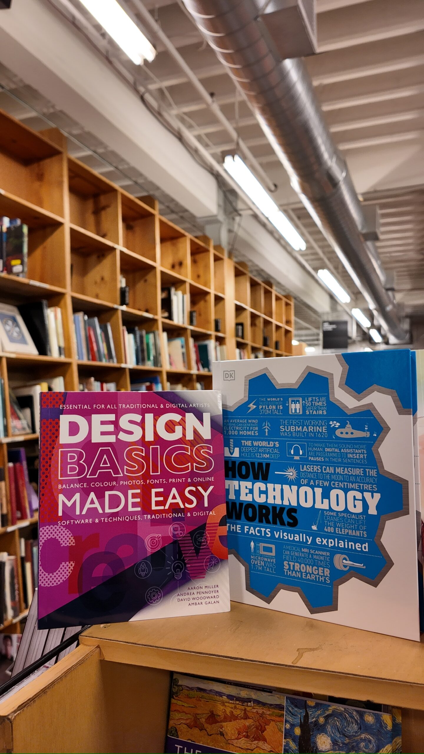 Two books, "Design Basics Made Easy" and "How Technology Works," are displayed upright on a wooden shelf in a bookstore with bookshelves in the background.