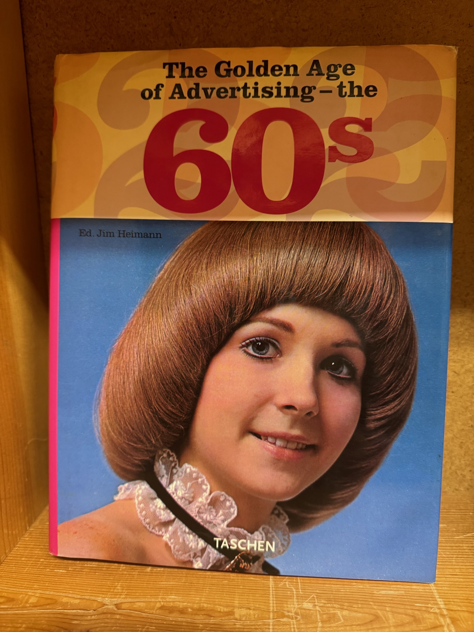 A book titled "The Golden Age of Advertising – the 60s" with a photo of a woman with a bob haircut and a white lace choker on the cover. Authored by Ed. Jim Heimann, published by Taschen.