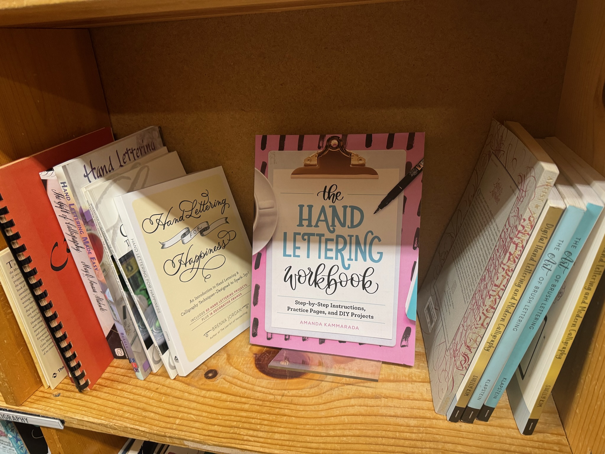 A selection of hand lettering books displayed on a wooden bookshelf, with "The Hand Lettering Workbook" prominently featured in the center.
