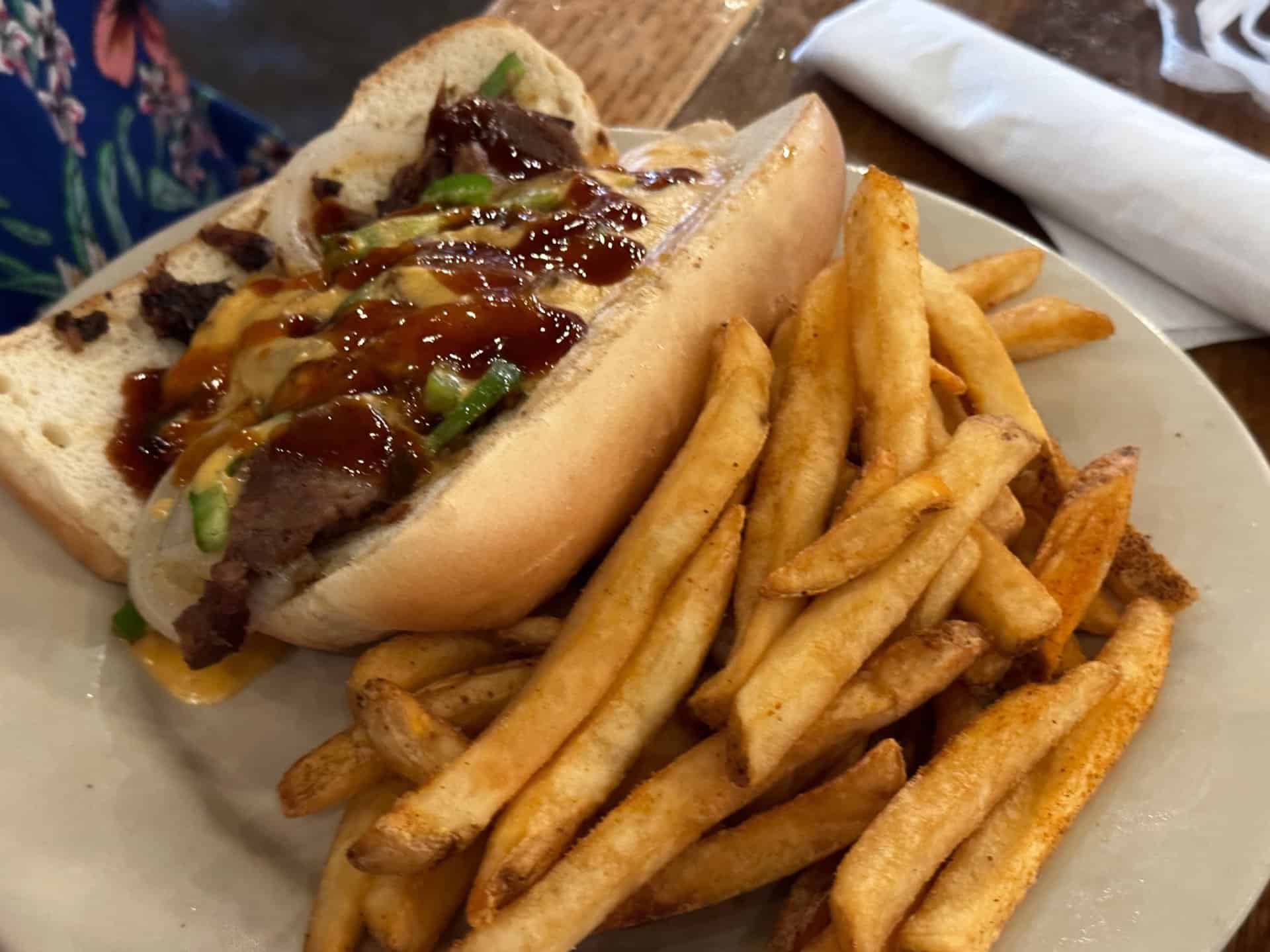 A Nashville-inspired sandwich with beef and sauce in a hoagie roll, served alongside seasoned fries on a white plate. Napkin in the background.