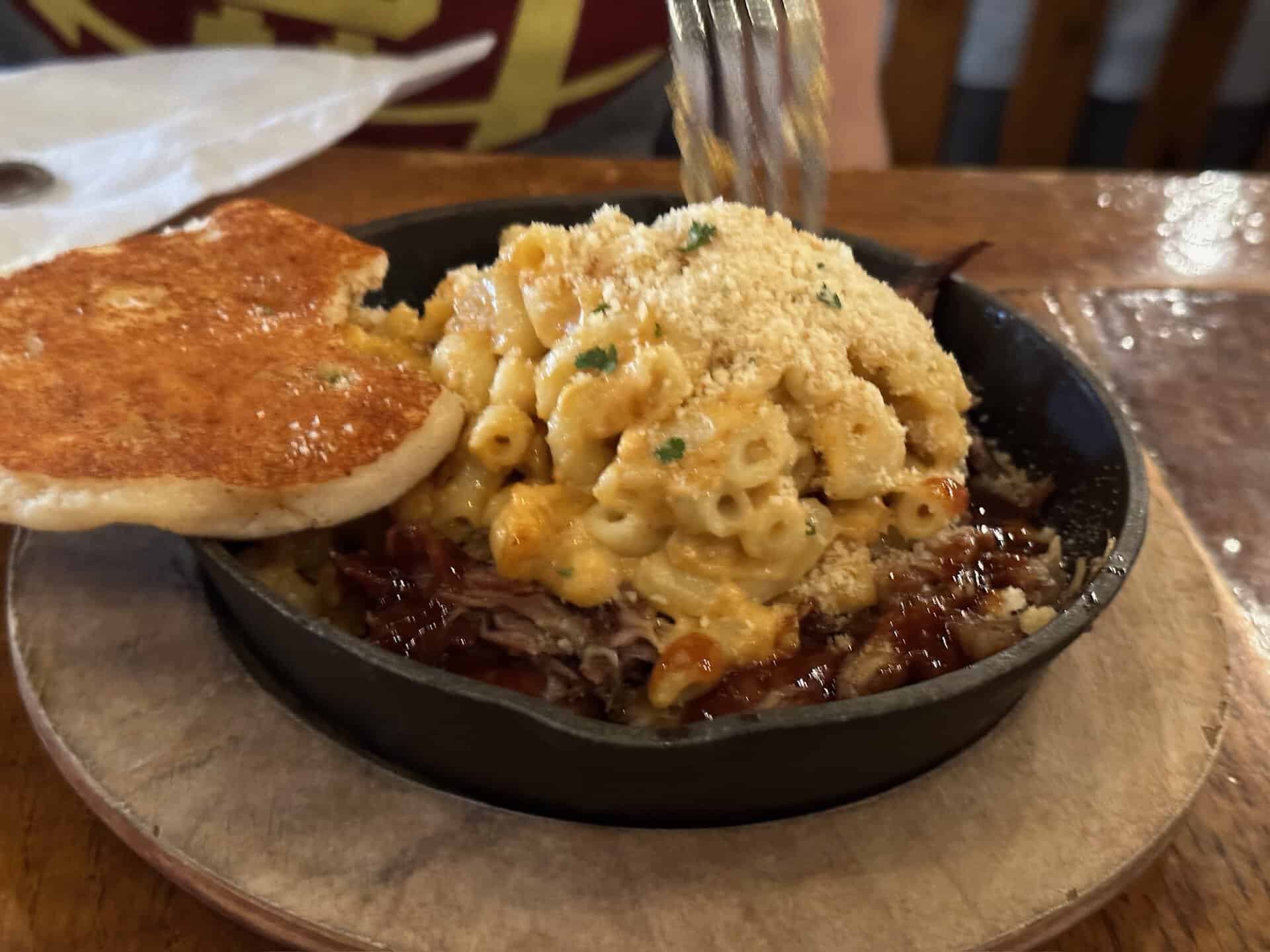 A Nashville twist on comfort food: a skillet of macaroni and cheese crowned with breadcrumbs, resting over shredded meat with BBQ sauce, served alongside toasted bread.
